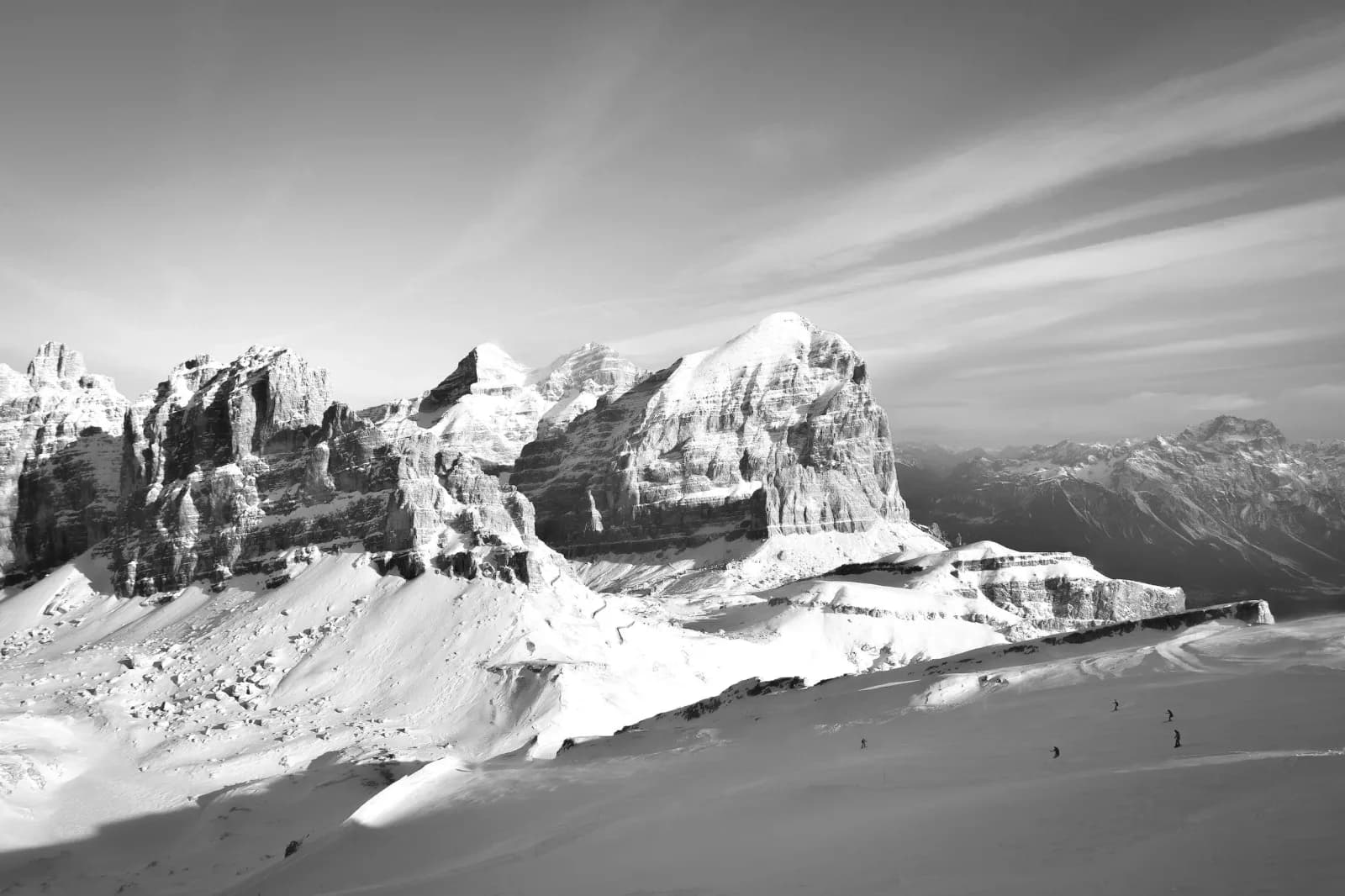 A black and white photograph captures a vast, snow-covered mountain landscape with dramatic rock formations and a cloudy sky.