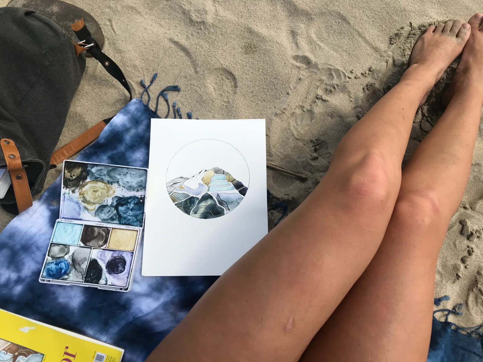 A watercolor painting of abstract mountains sits on the sand next to a palette of paints.