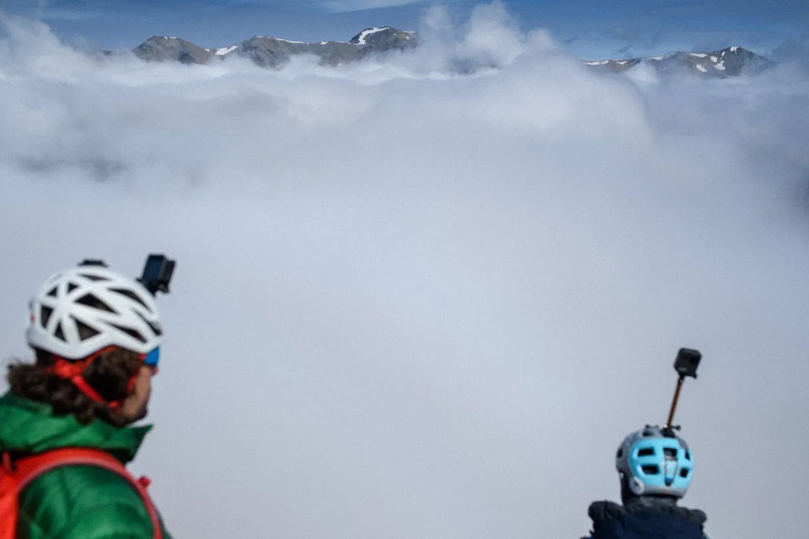 Two skiers in helmets with action cameras stand above the clouds with mountains in the distance.