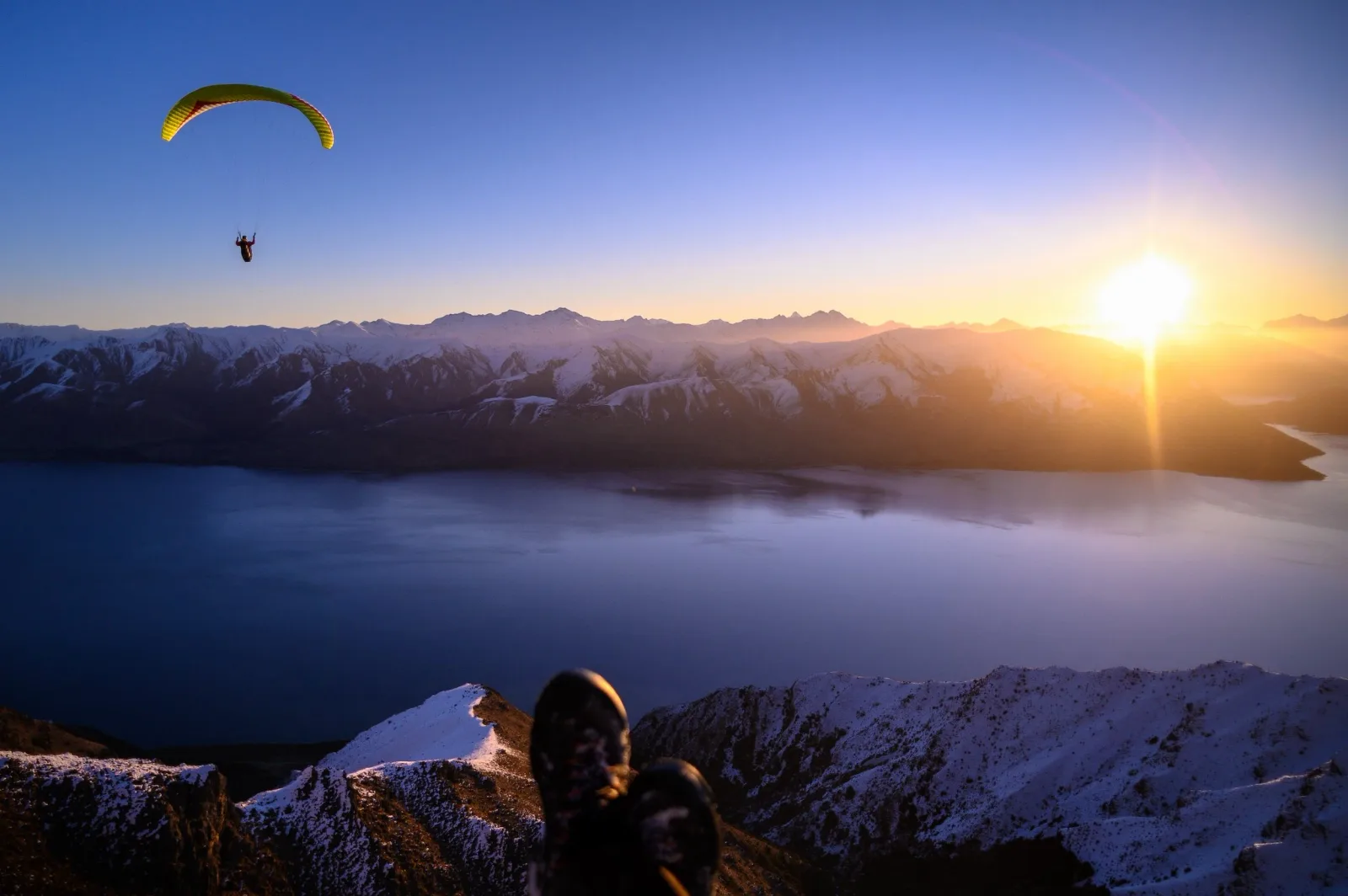 A paraglider soars over a snow-capped mountain range and a deep blue lake at sunset.