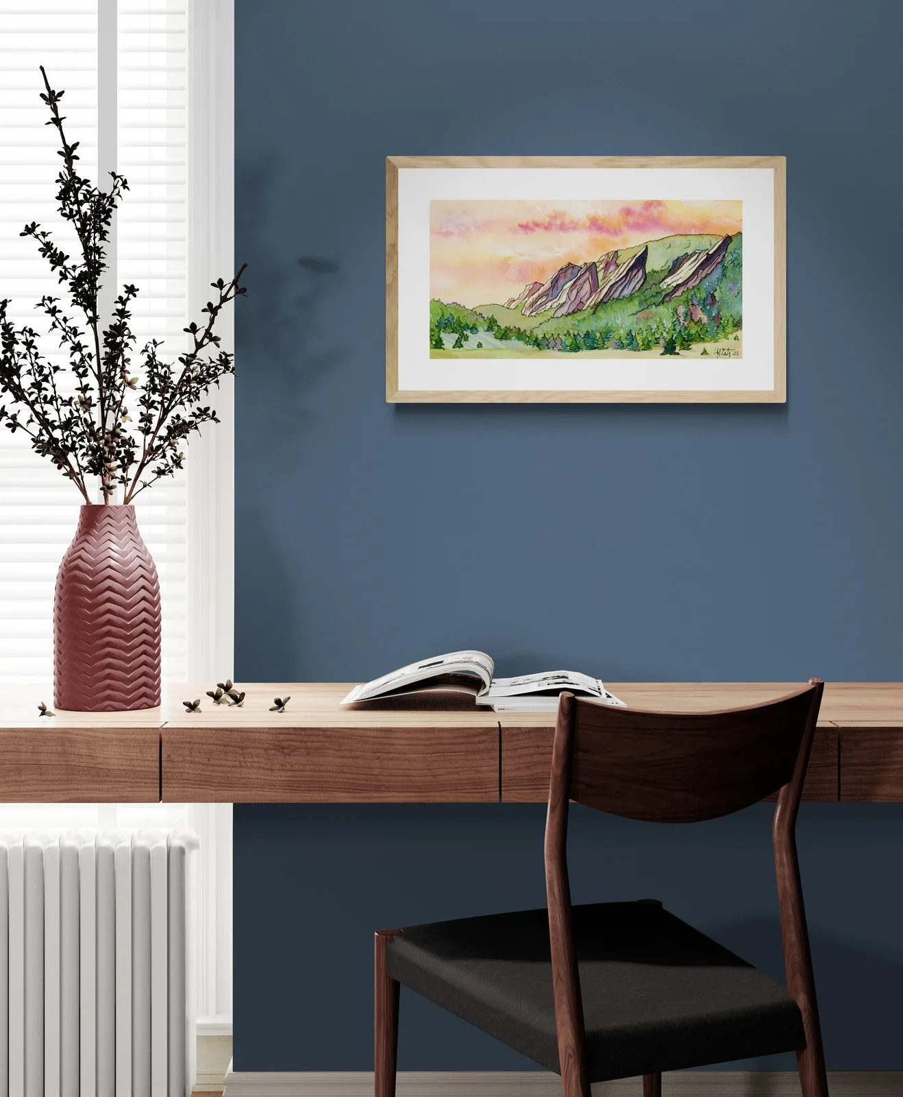 A watercolor painting of the Flatirons mountains at sunset hangs above a desk in a furnished room.