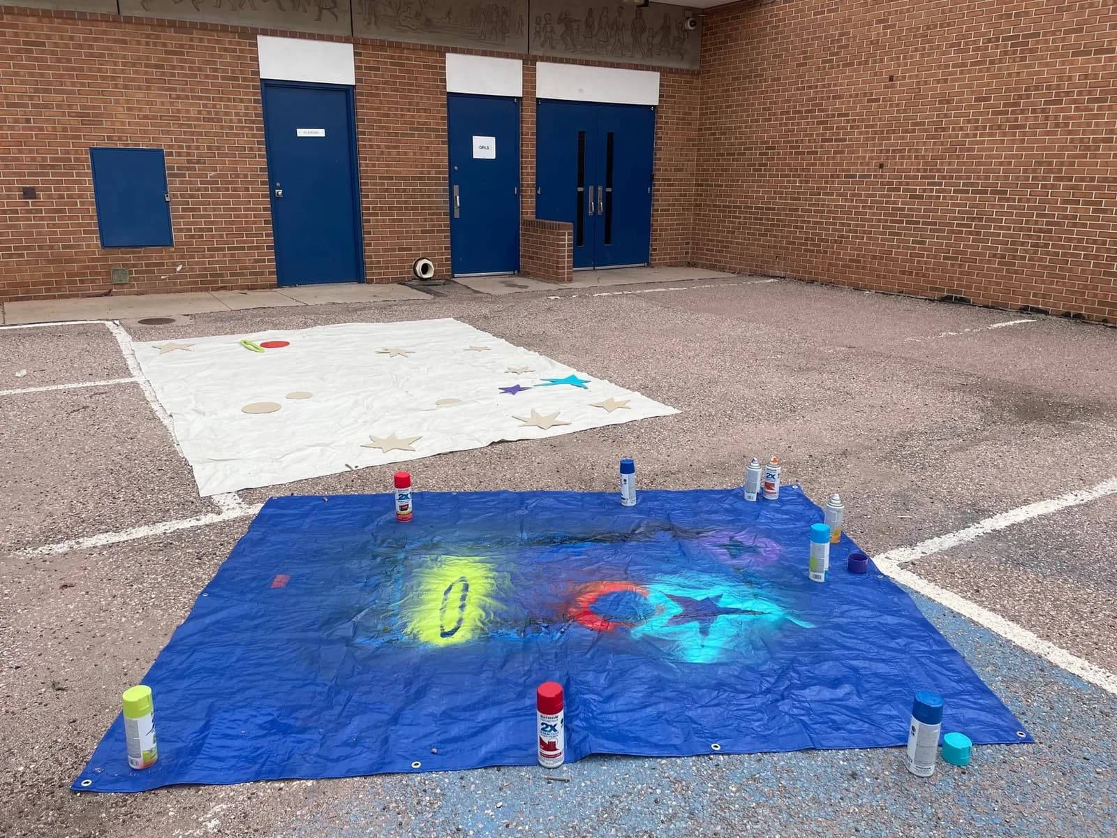 Spray paint art is being created on a blue tarp in an outdoor setting with a brick building in the background.