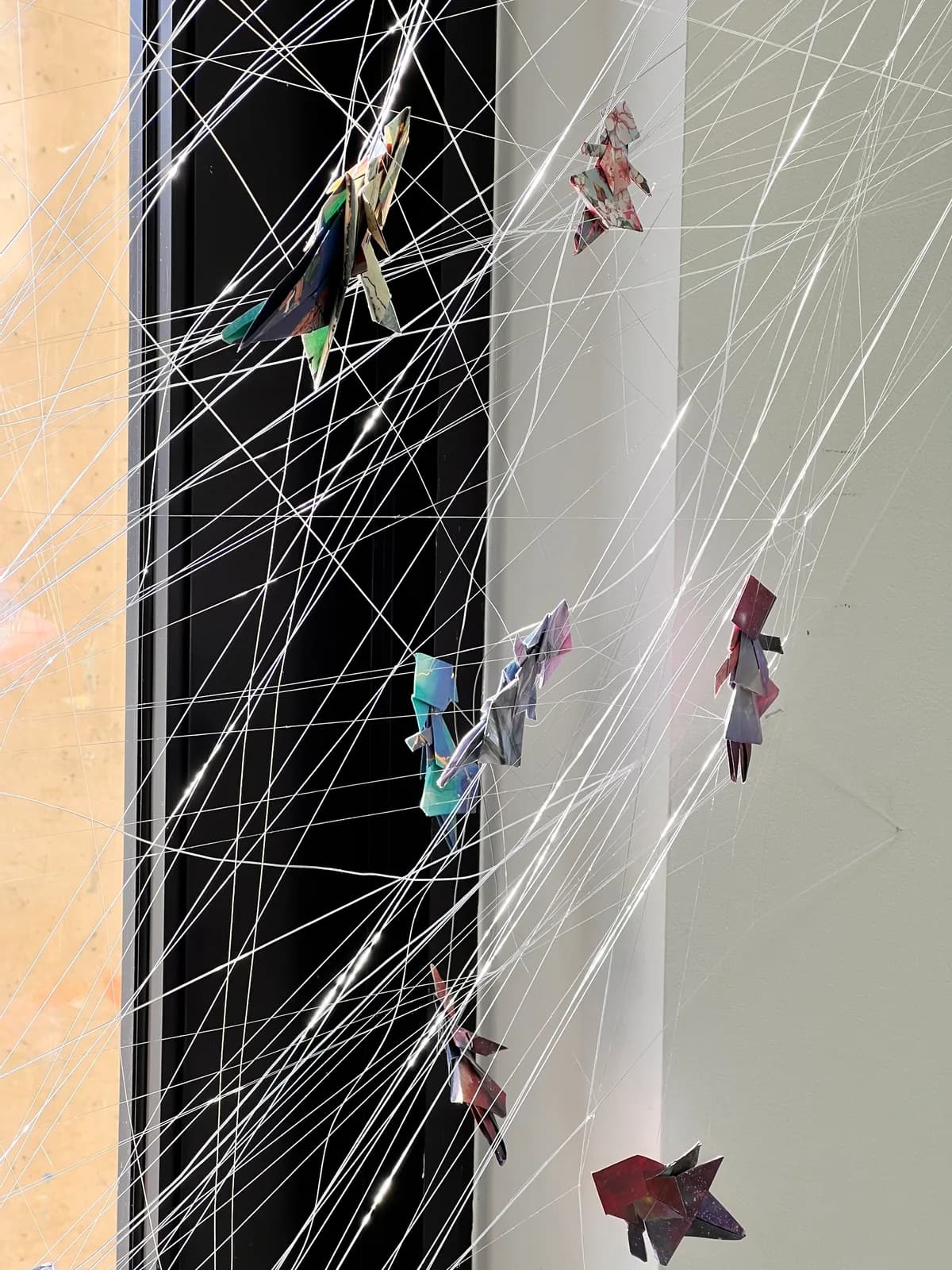 An abstract mixed media installation features numerous folded paper figures suspended by thin white threads against a dark background.
