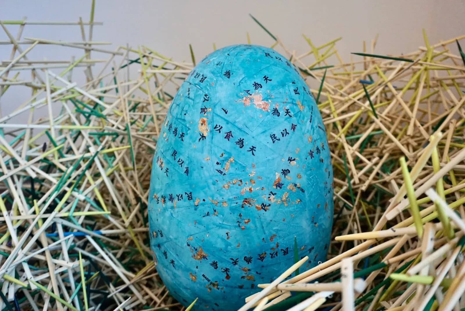 A mixed media sculpture of a turquoise egg with gold leaf and Asian characters, nestled in a chaotic nest of bamboo.