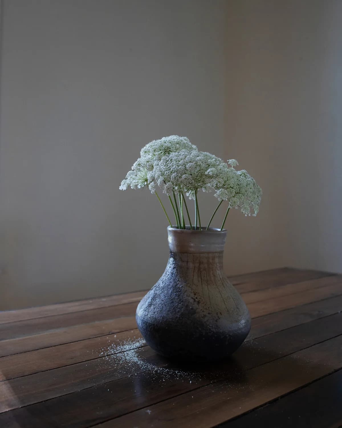 A ceramic vase holds delicate white flowers, set on a wooden table with scattered dust.
