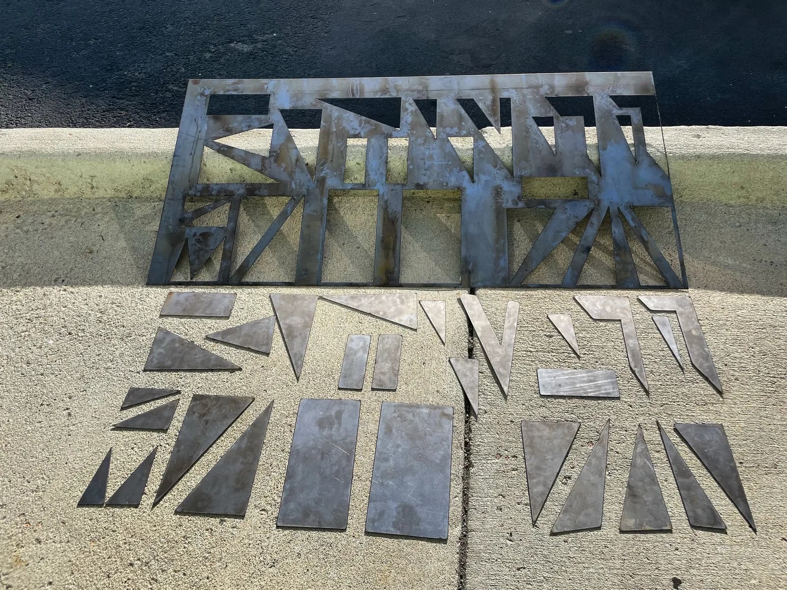 Metal geometric shapes are laid out on a concrete surface, suggesting an abstract sculpture in progress.