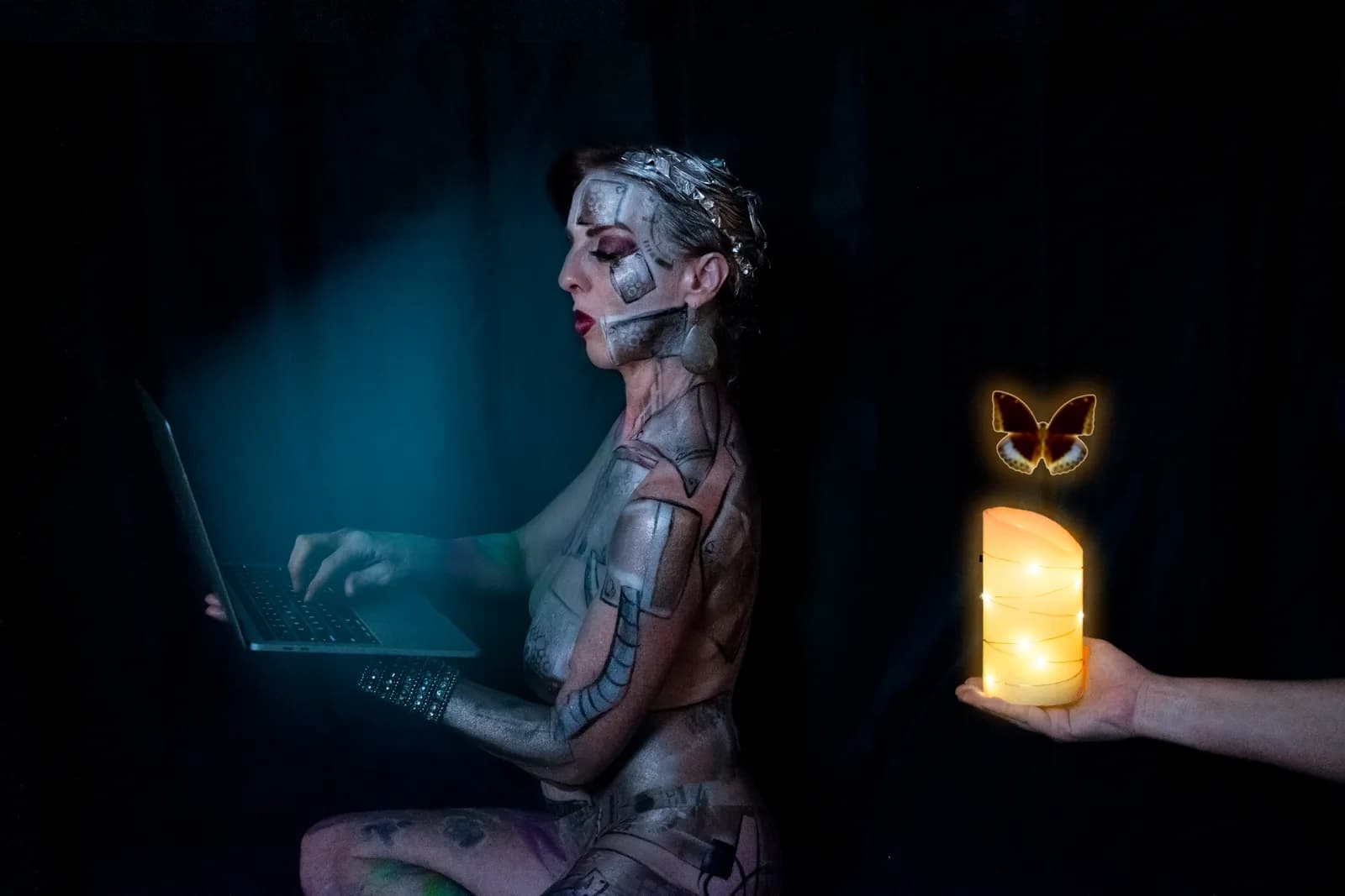 A digitally altered photograph features a figure with robot-like body paint interacting with a laptop, beside a candle and butterfly.