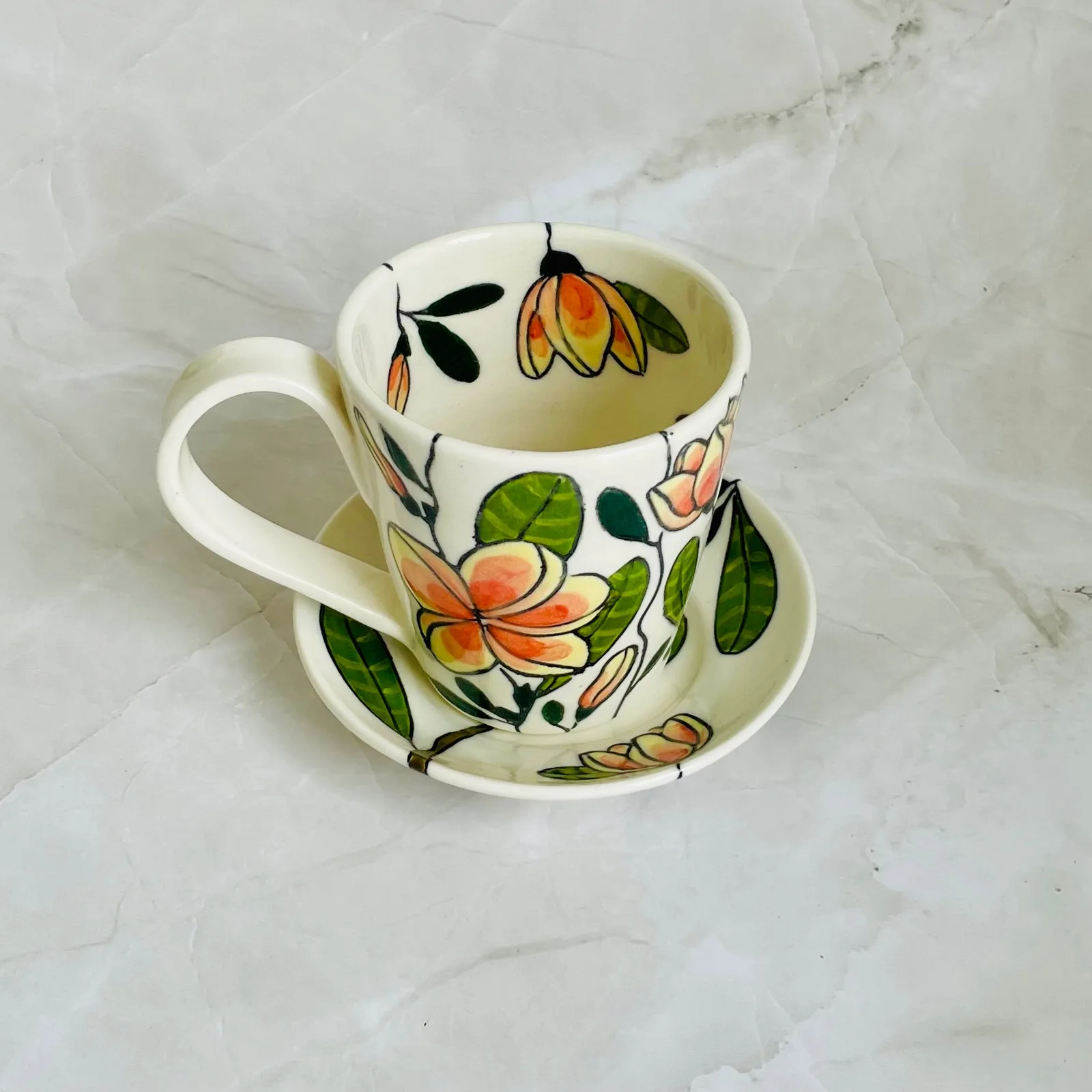 A hand-painted ceramic cup and saucer feature a floral design in Tuscany and Pumice hues.