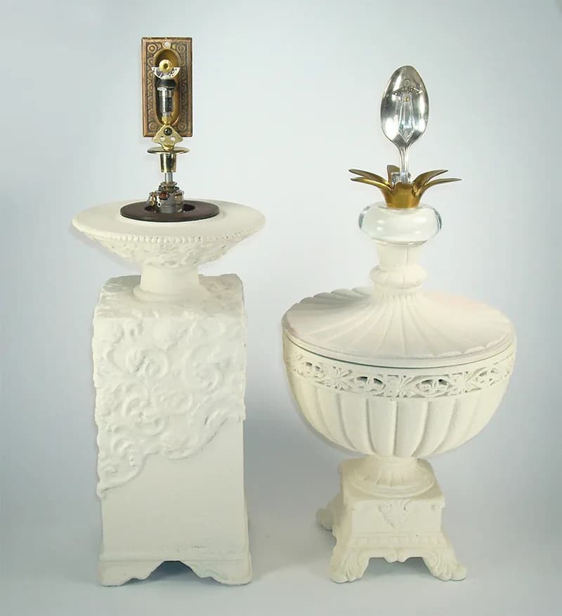 Two mixed media sculptures on pedestals, one topped with a spoon and the other with hardware, in a matte white finish.