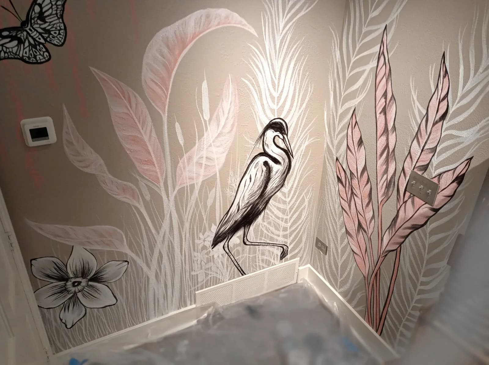 A black ink drawing of a heron stands amidst stylized foliage and a butterfly, rendered on a sandstone-colored wall.