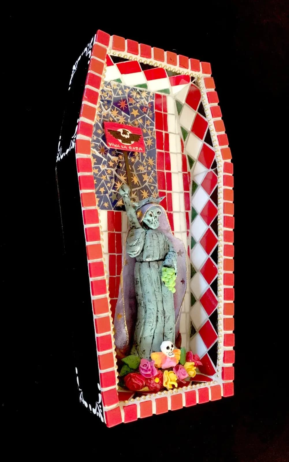 A mosaic-encased ceramic figure of a skeleton holding grapes, adorned with flowers and a flag.