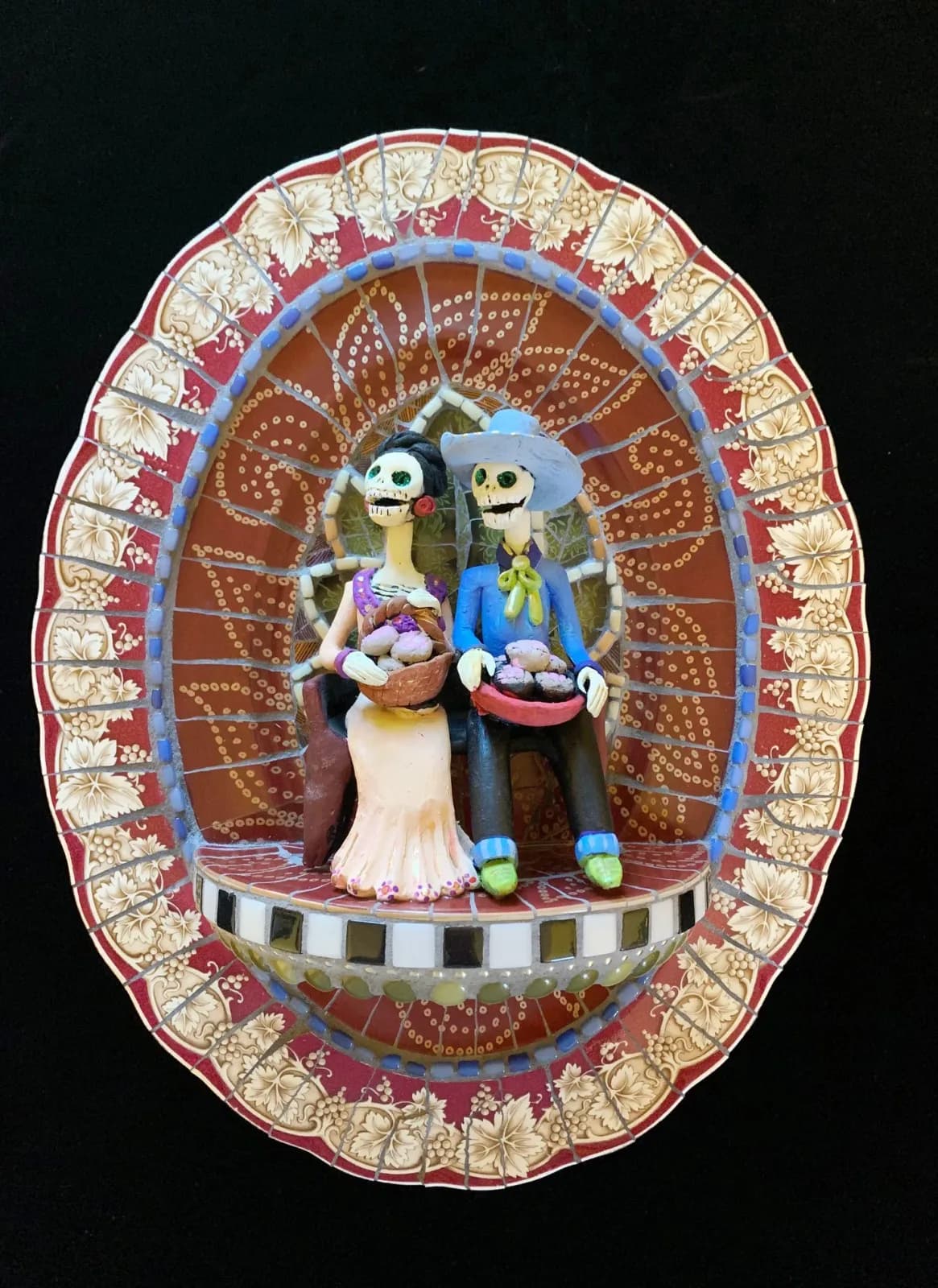 A folk art mosaic sculpture depicts two skeleton figures holding baskets of produce, rendered in earthy tones.