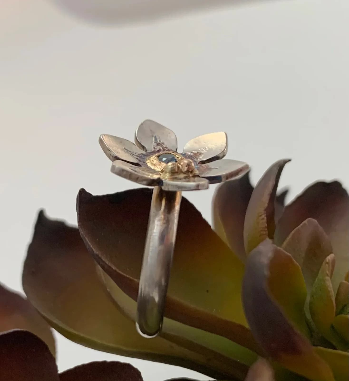 A silver and gold-accented floral ring with a small gemstone, crafted by a metalsmith.