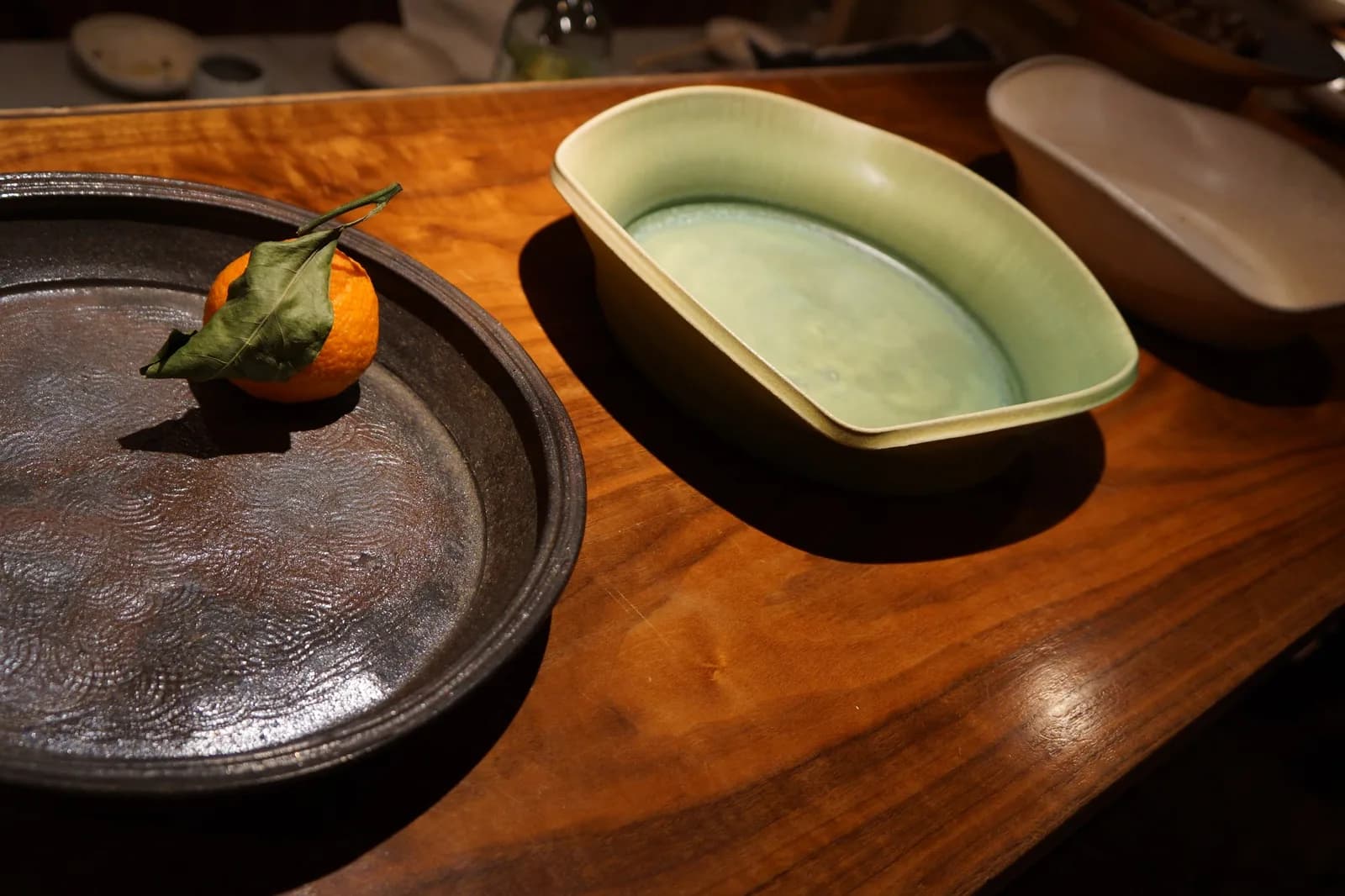 A dark ceramic dish holds an orange with a leaf, next to a pale green ceramic vessel on a wooden surface.