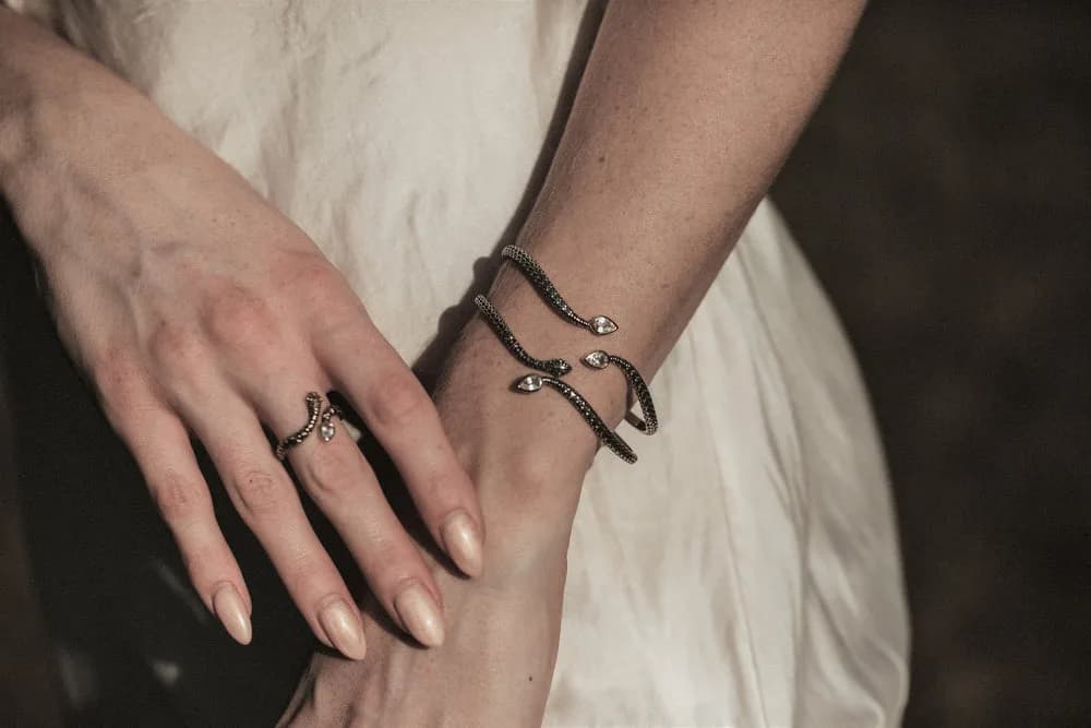 A metal snake bracelet and ring adorn a person's arm and hand, set against a neutral background.