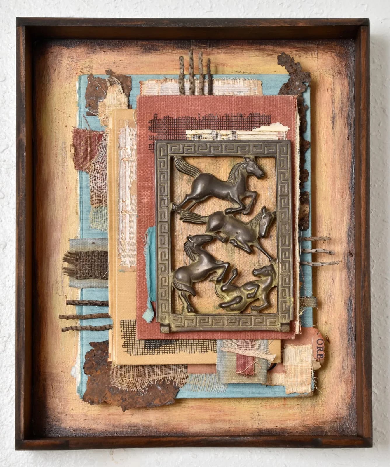 A mixed media assemblage featuring metal horses within a rustic frame, incorporating fabric and paper elements.