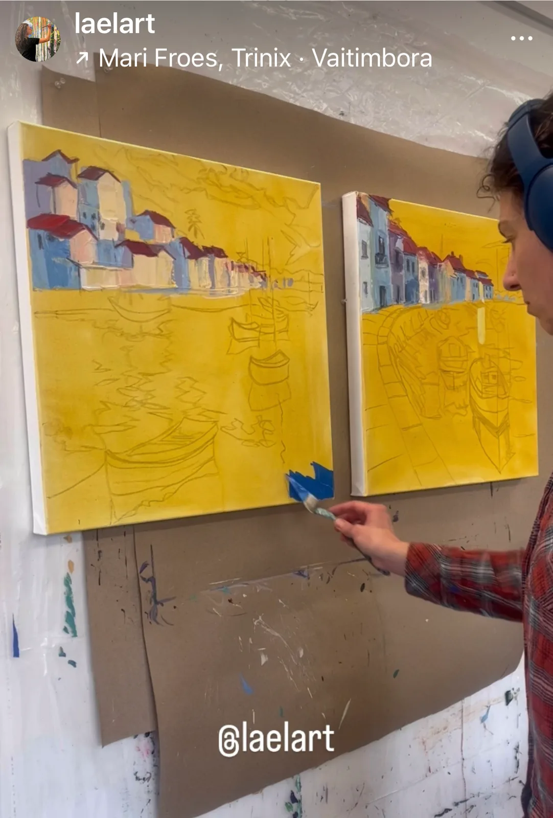 Artist painting two canvases in a studio with a bright yellow underpainting and a coastal village scene.