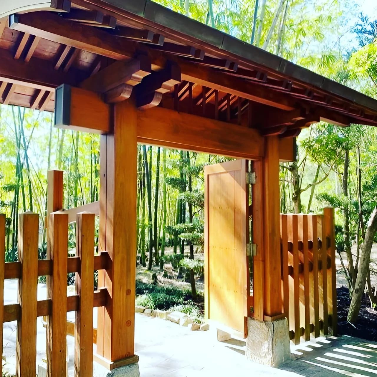 A wooden gate opens to a serene garden with tall bamboo and manicured trees, set in a natural landscape.