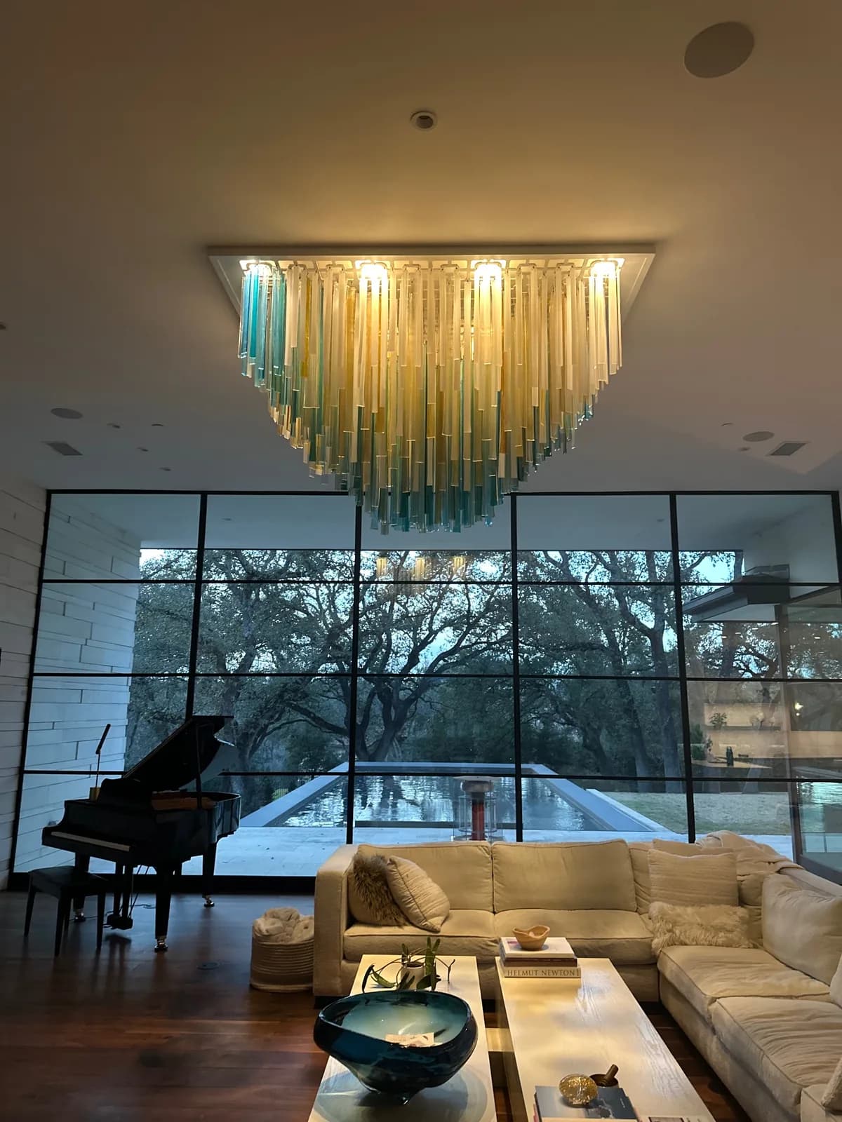 A glass chandelier hangs above a living room with a piano, sofa, and view of a pool and trees.