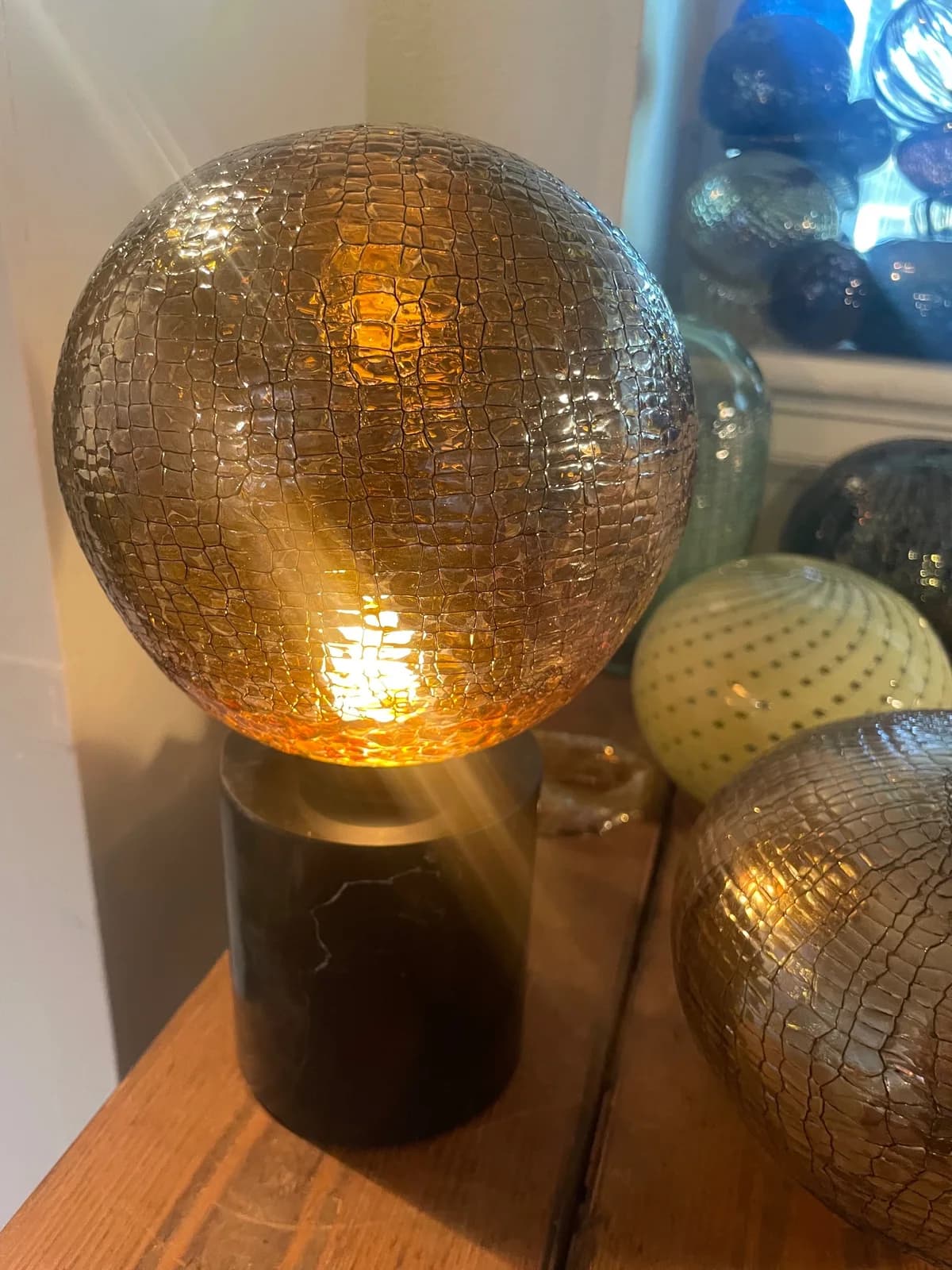 A textured glass orb lamp with a dark base sits on a wooden table with other decorative glass objects.