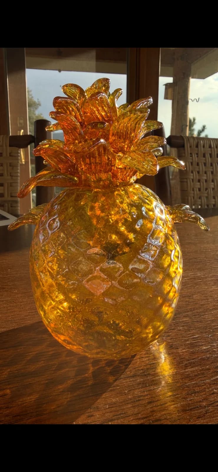 A glass pineapple sculpture sits on a wooden table, with a blurred outdoor scene visible in the background.