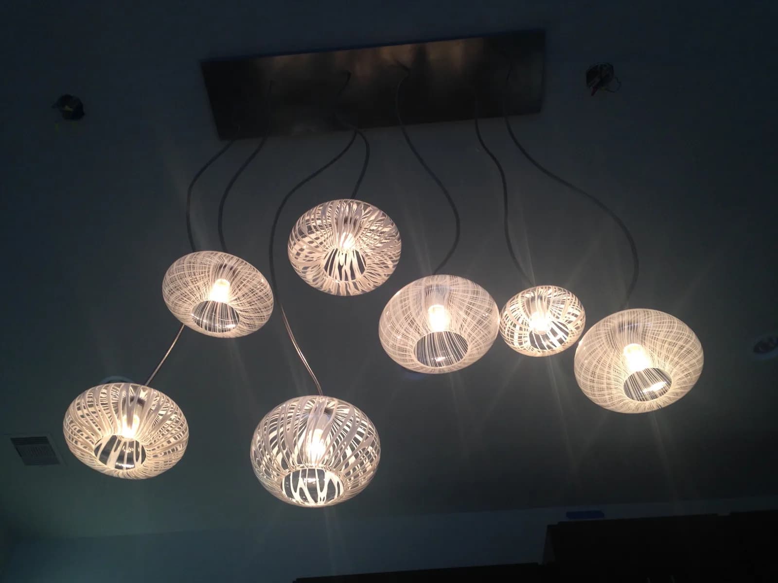 A cluster of seven pendant lights with white woven shades hang from a dark ceiling.
