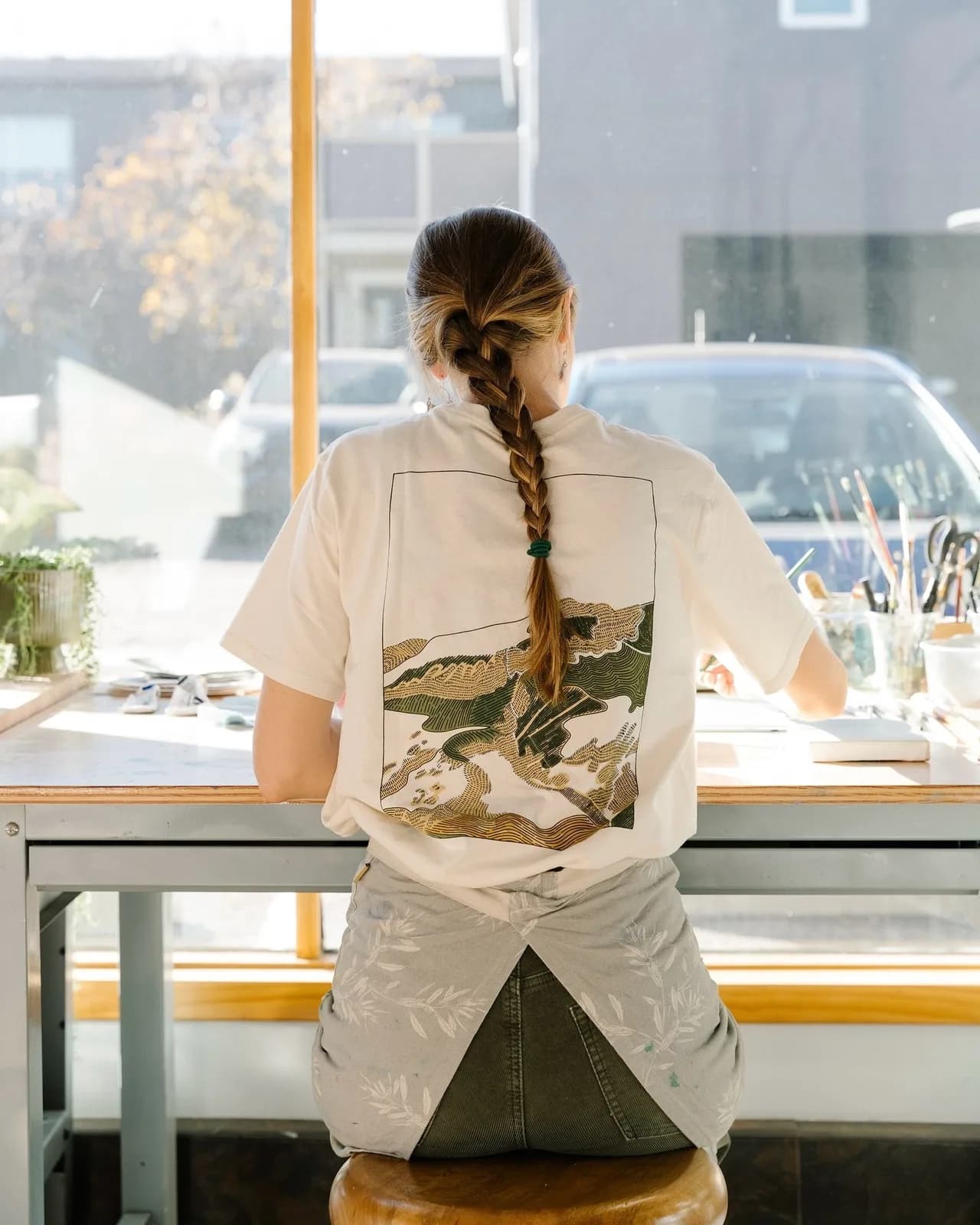 A person wearing a t-shirt with a landscape design sits at a table in a studio with art supplies.