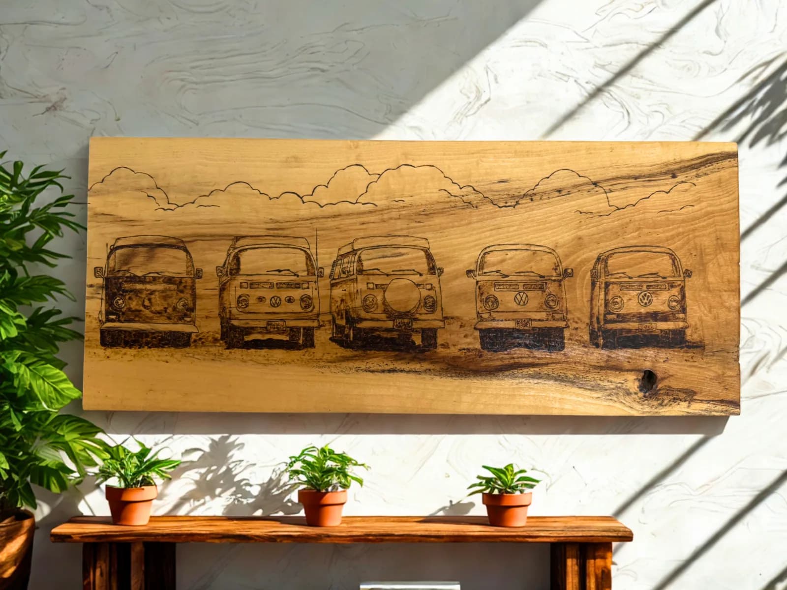 A wood-burned artwork depicts five vintage Volkswagen buses lined up under a cloudy sky.