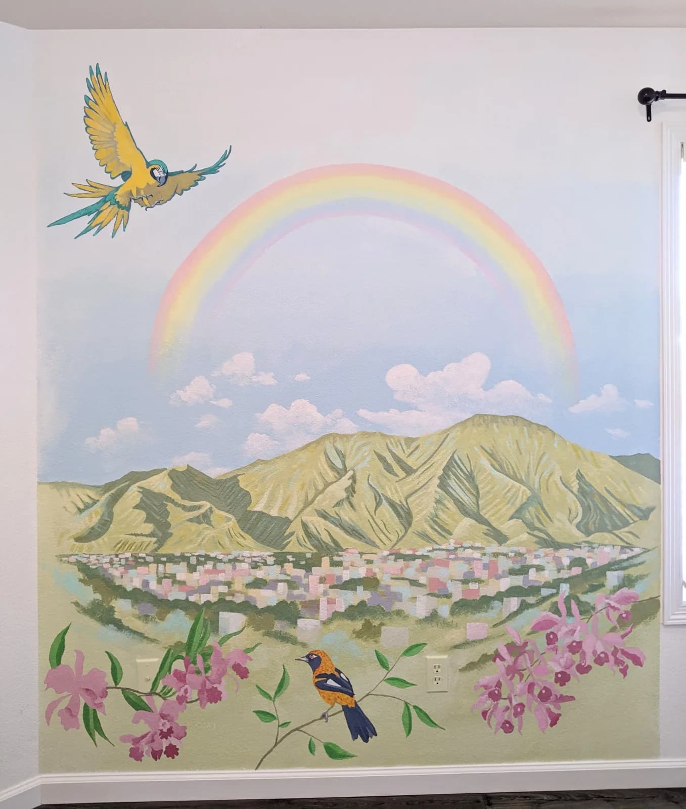 A vibrant mural depicts a cityscape nestled below mountains, with a parrot flying near a rainbow and birds perched on flowering branches.