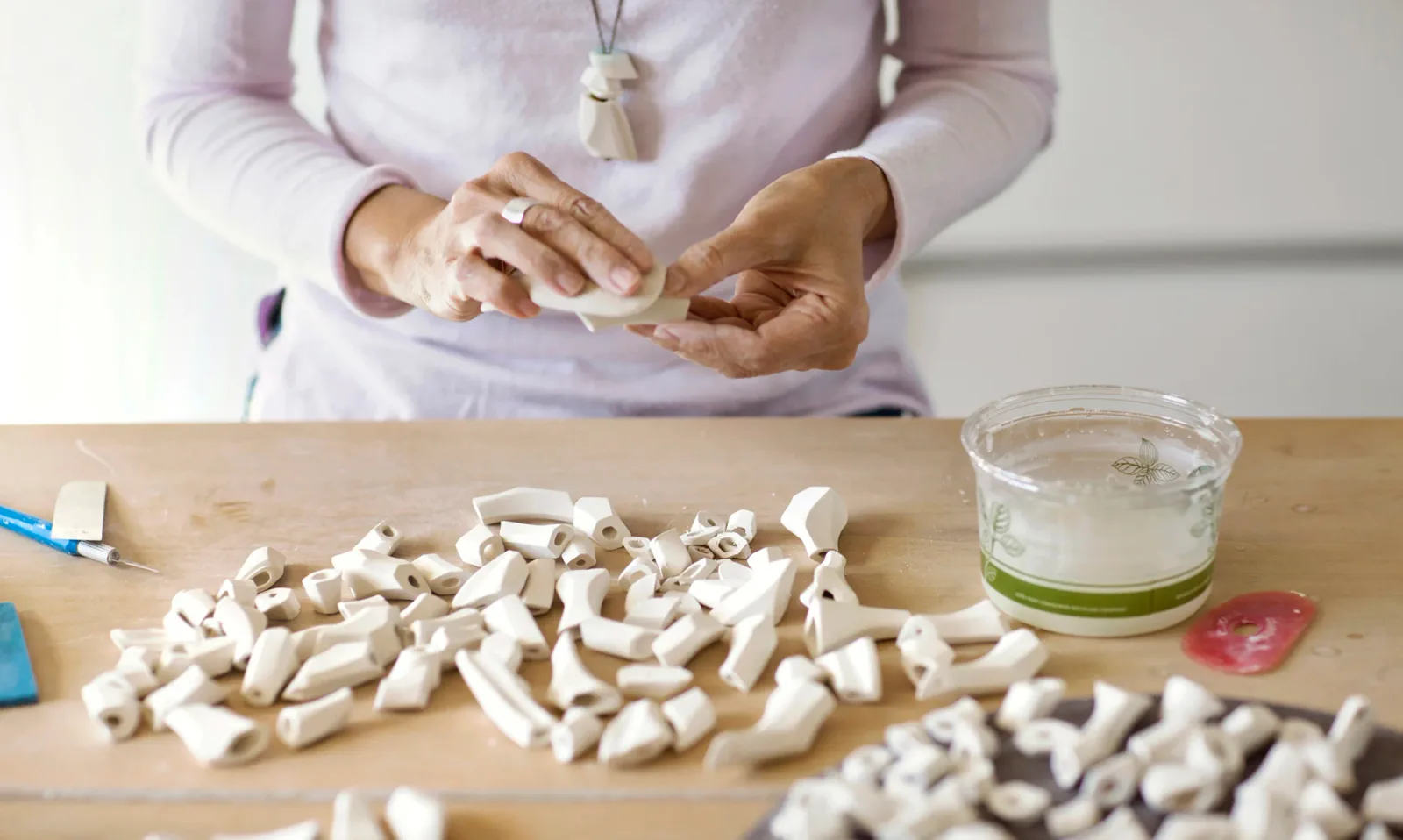 An artist's hands work with clay pieces on a wooden table, surrounded by many small ceramic components.
