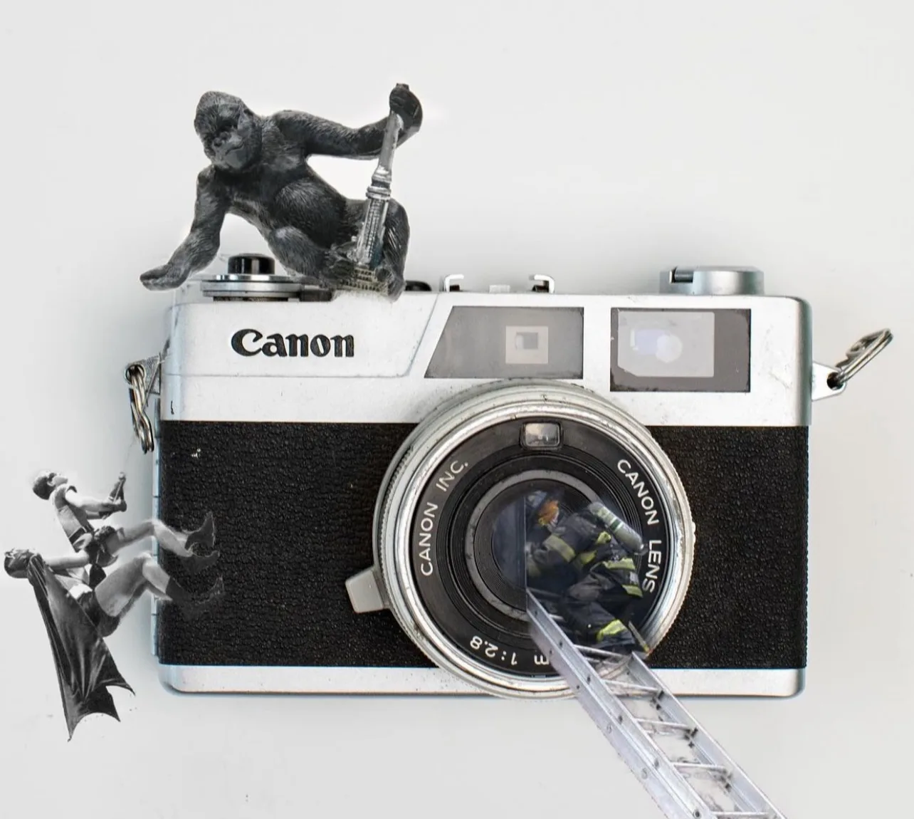 A surreal scene depicts a toy gorilla and falling superheroes around a vintage Canon camera, with a firefighter climbing a ladder into the lens.