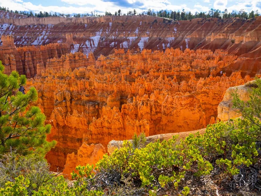 A vibrant landscape photograph captures the orange hoodoos of Bryce Canyon under a bright sky with scattered snow.