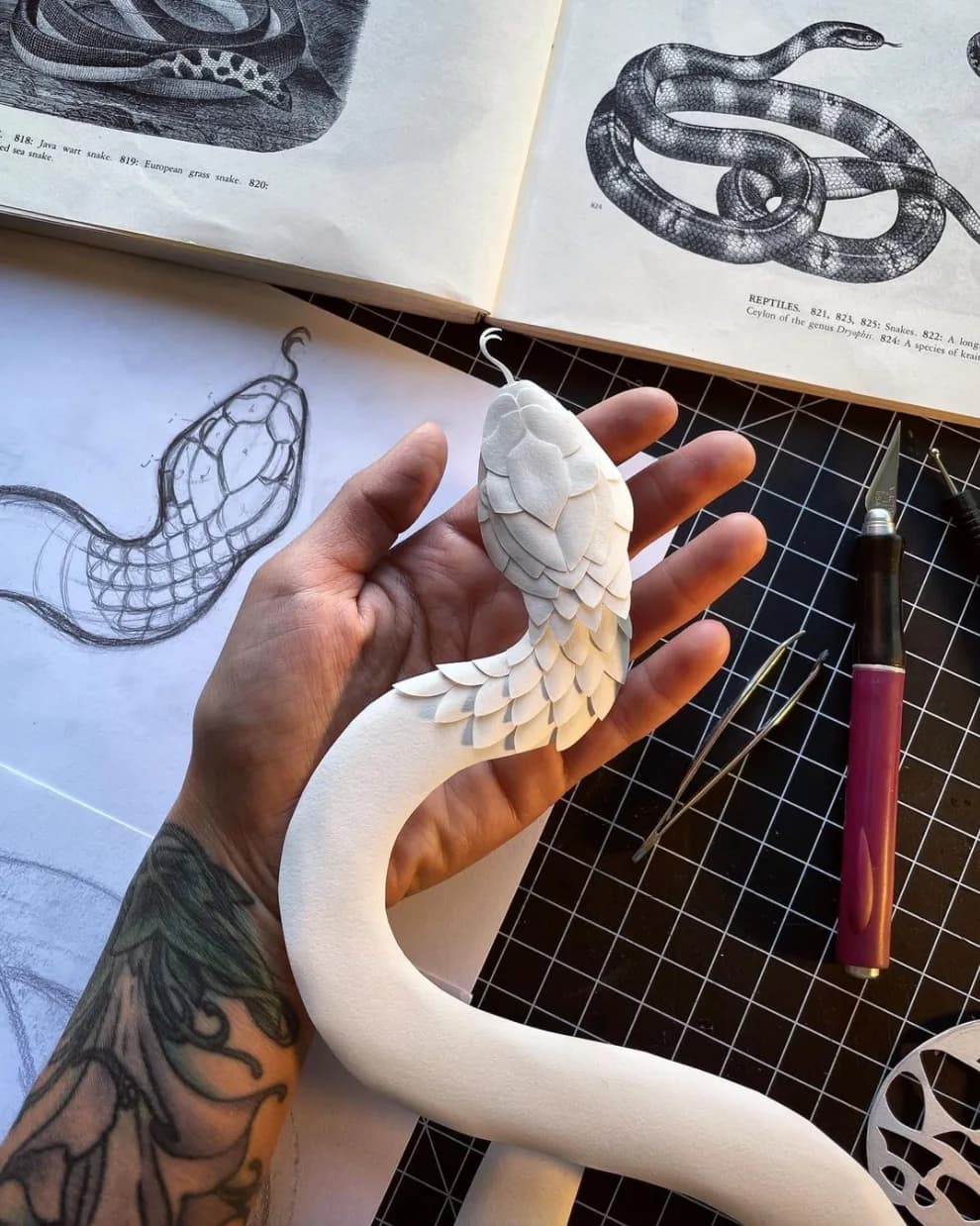 A white paper sculpture of a snake is held in a hand, with reference images and tools visible.