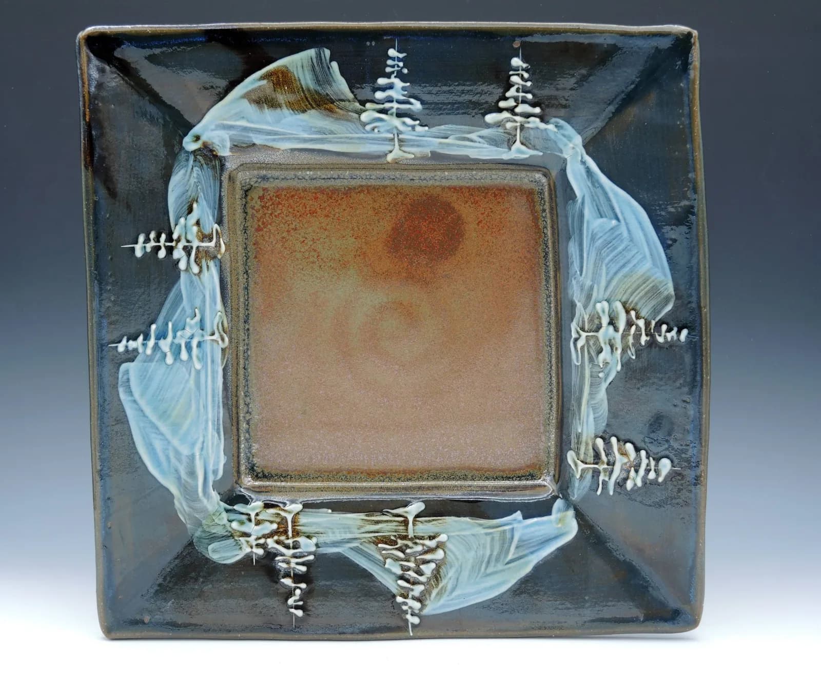 A square ceramic plate features a winter landscape with stylized trees and a warm, earthy central glaze.