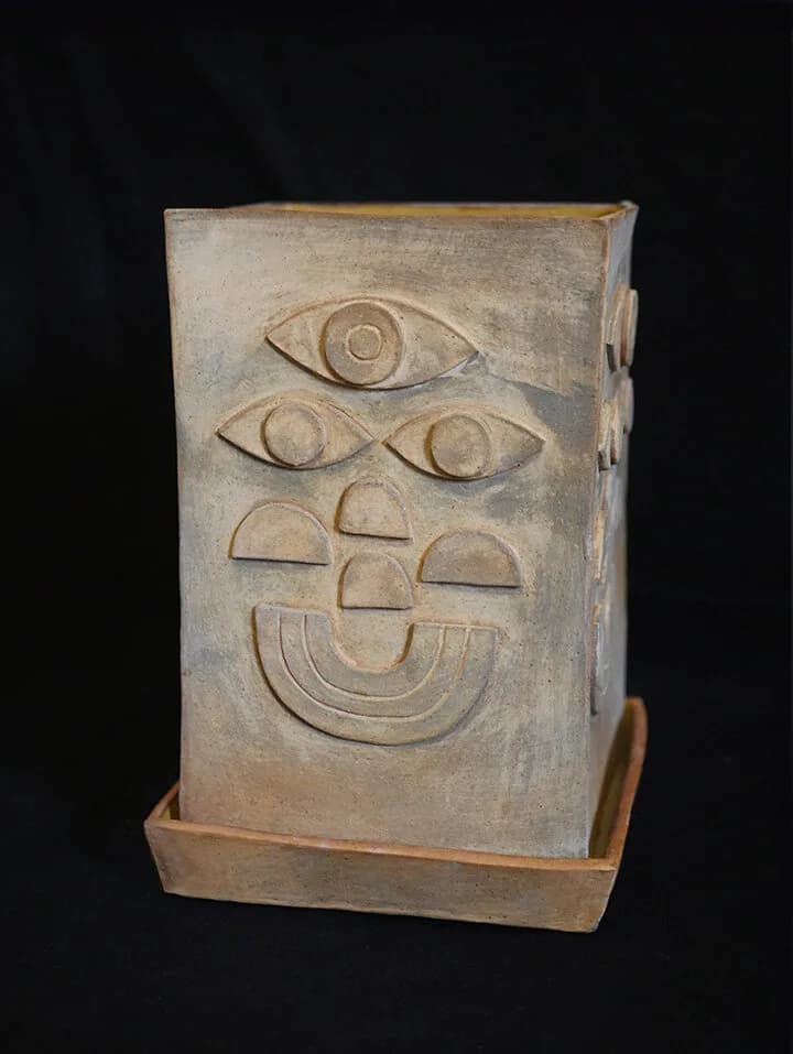 A ceramic sculpture with a stylized face, featuring multiple eyes and a smiling mouth, in shades of Stonewall and Cement.