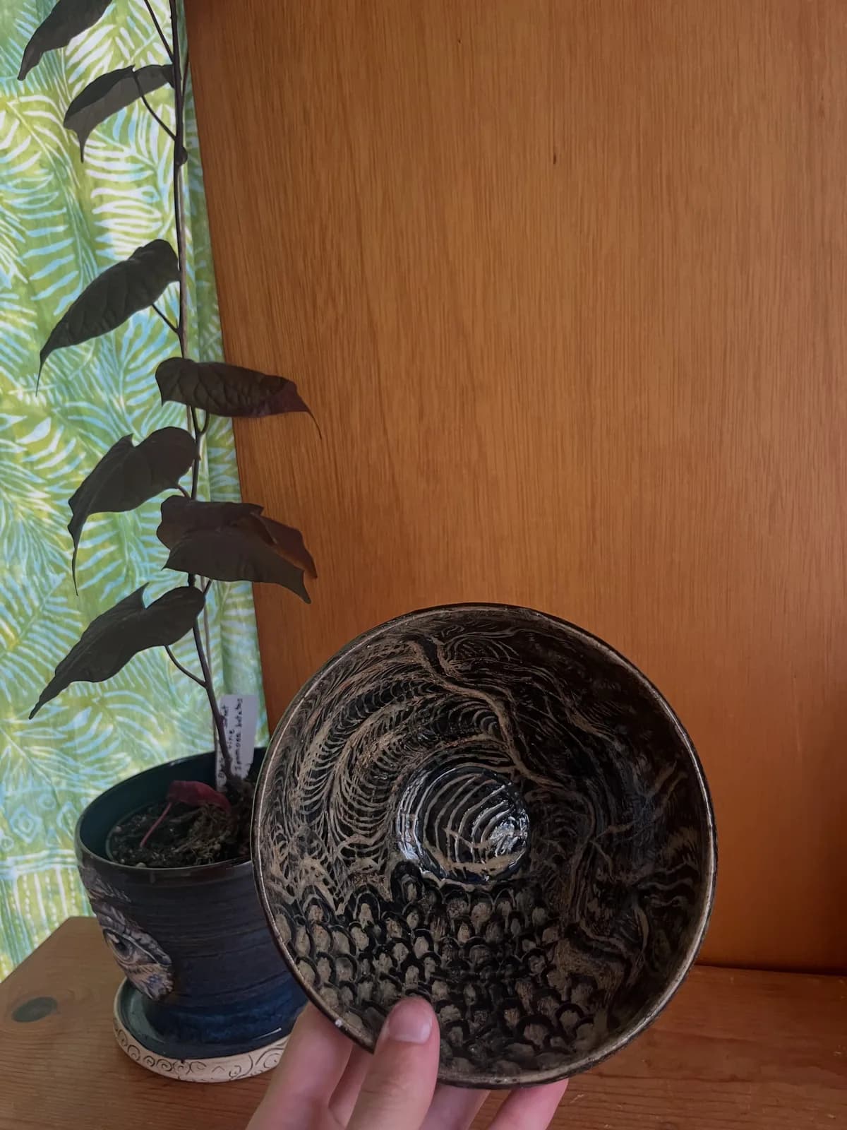 A hand-decorated ceramic bowl is shown in situ next to a potted plant with dark leaves and a patterned curtain.