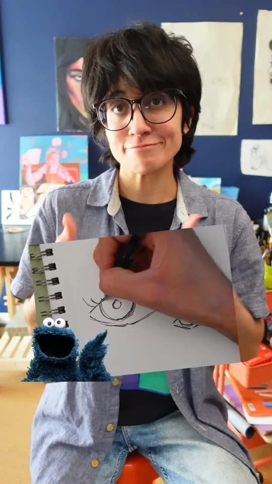 A person sketches an eye in a sketchbook, with artwork visible on the wall behind them.