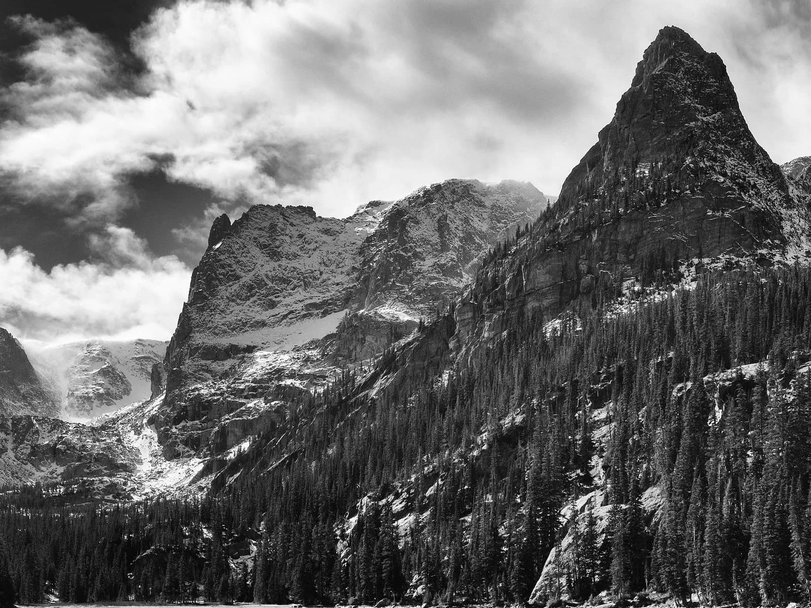 A black and white photograph captures a dramatic mountain landscape with dense evergreen trees and dramatic clouds.