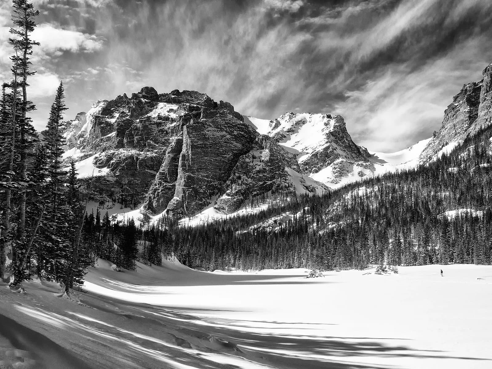 A black and white photograph captures a vast winter landscape with snow-covered mountains, pine trees, and a frozen lake.