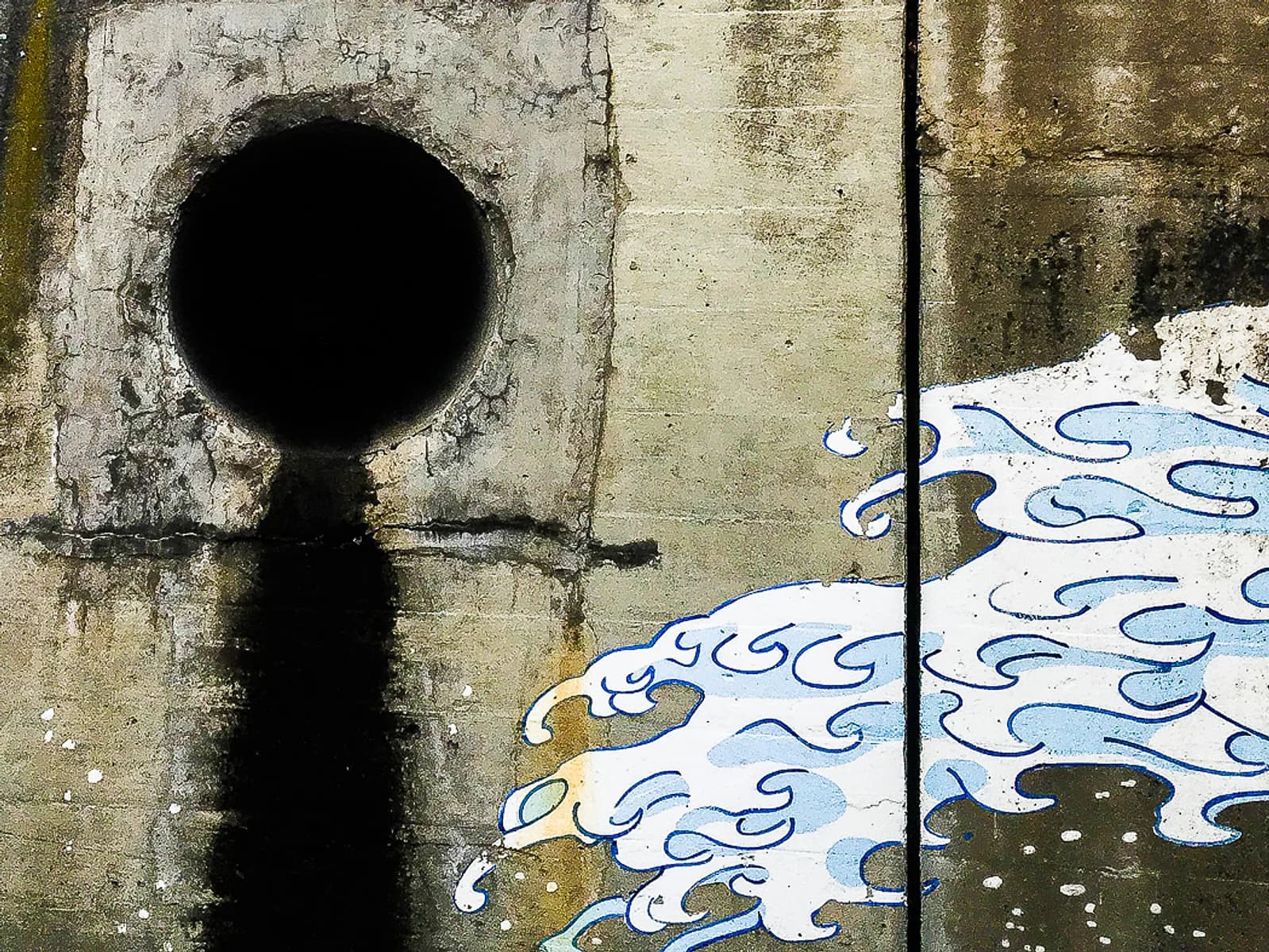 A street art mural depicting stylized waves painted on a concrete wall with a drainage pipe.