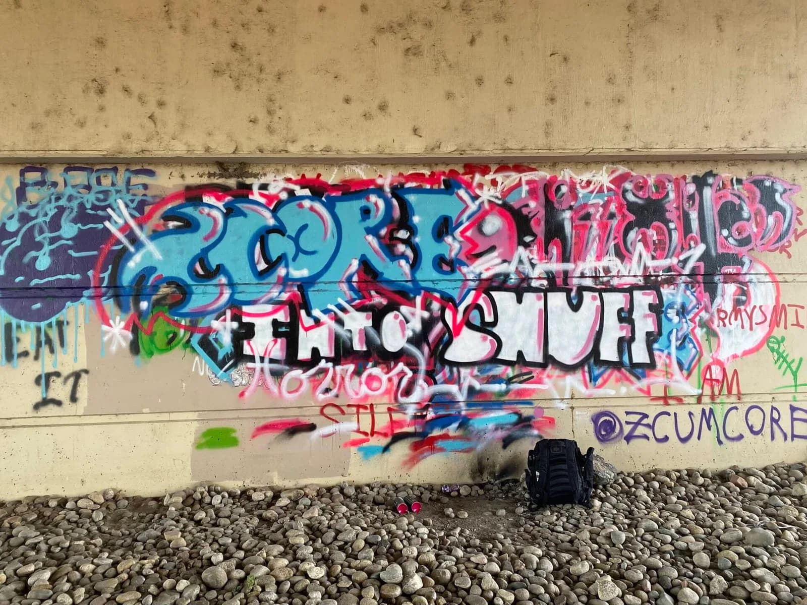 A vibrant graffiti mural covers a concrete wall, with a backpack resting on the gravel below.