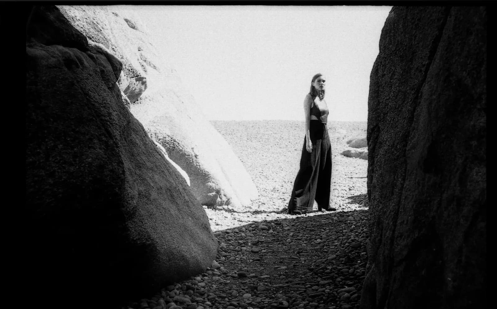 A black and white photograph captures a woman standing between large rocks on a pebble beach.
