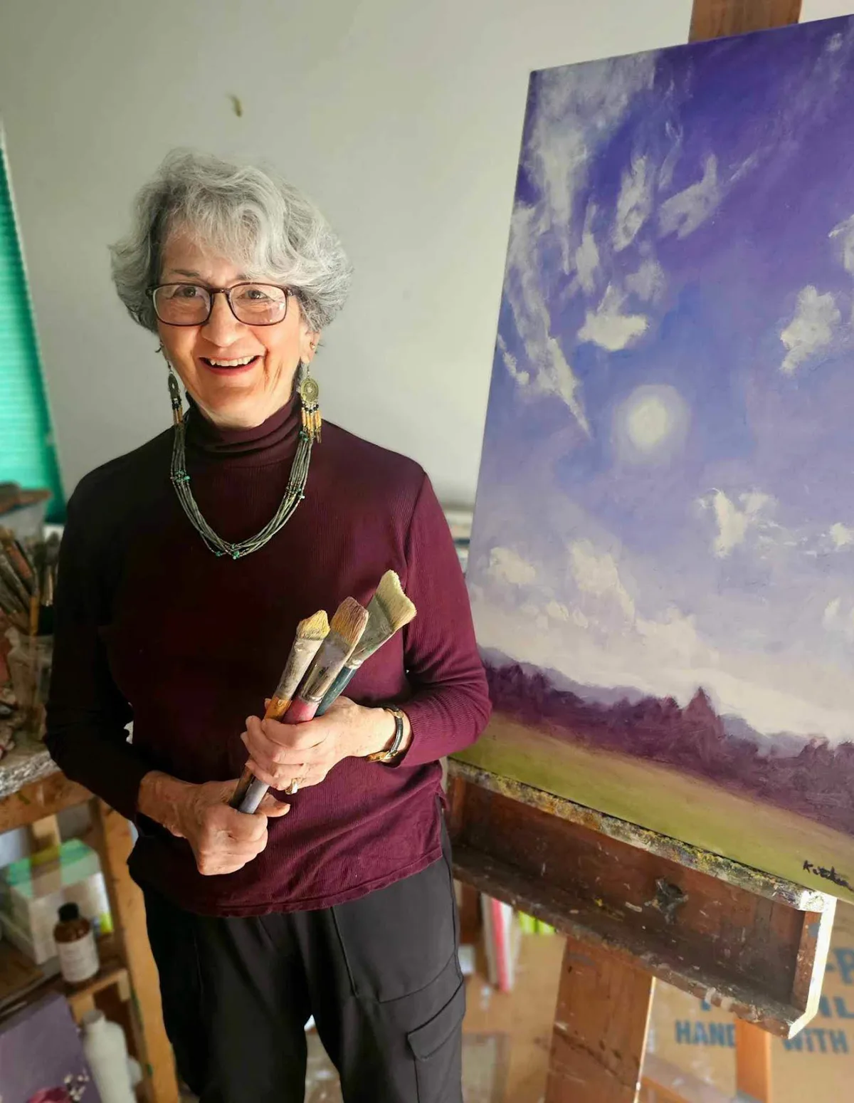 An artist holds brushes next to an impressionistic landscape painting in a studio.