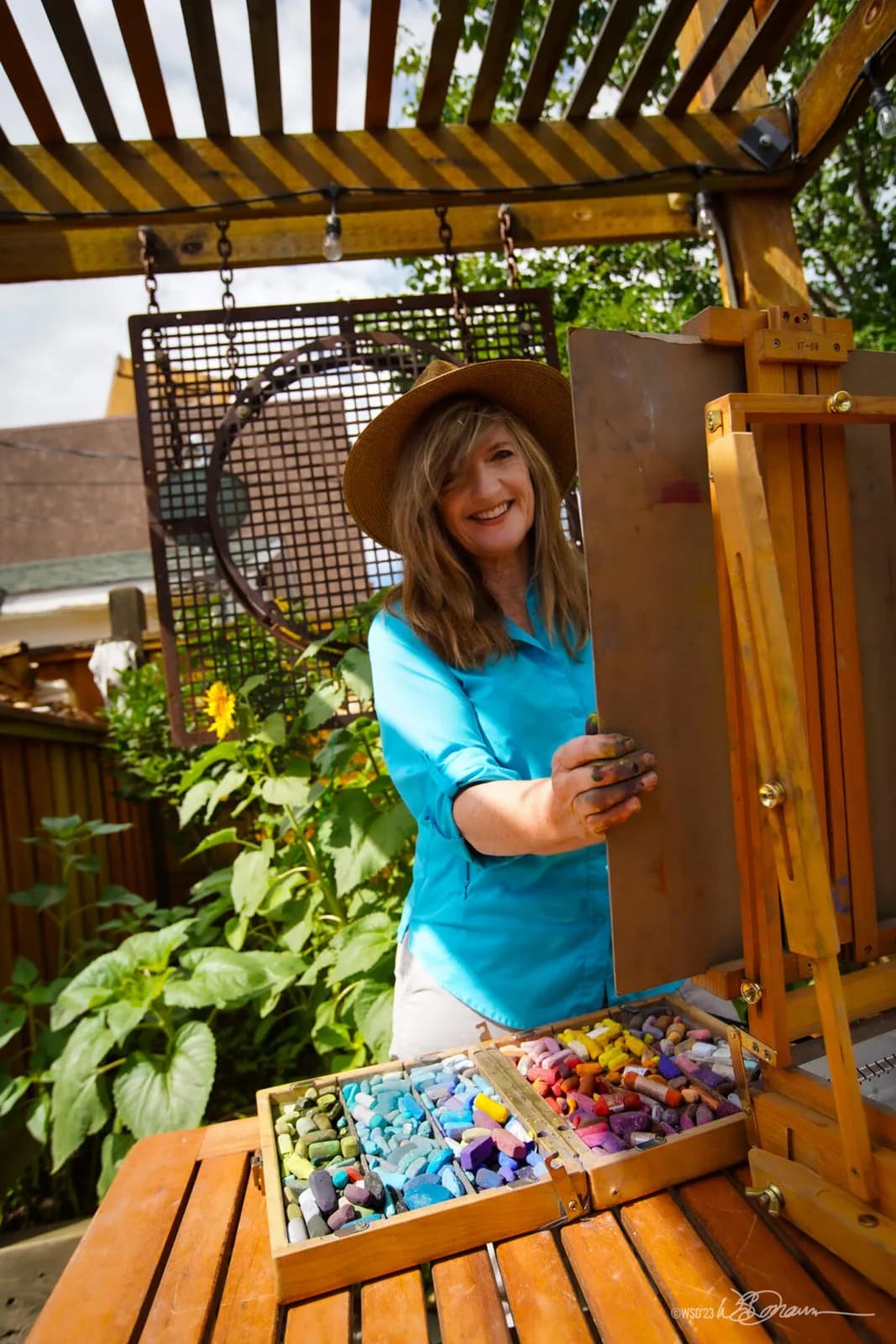 An artist in a straw hat smiles while holding a board in front of an easel with a box of colorful pastels.