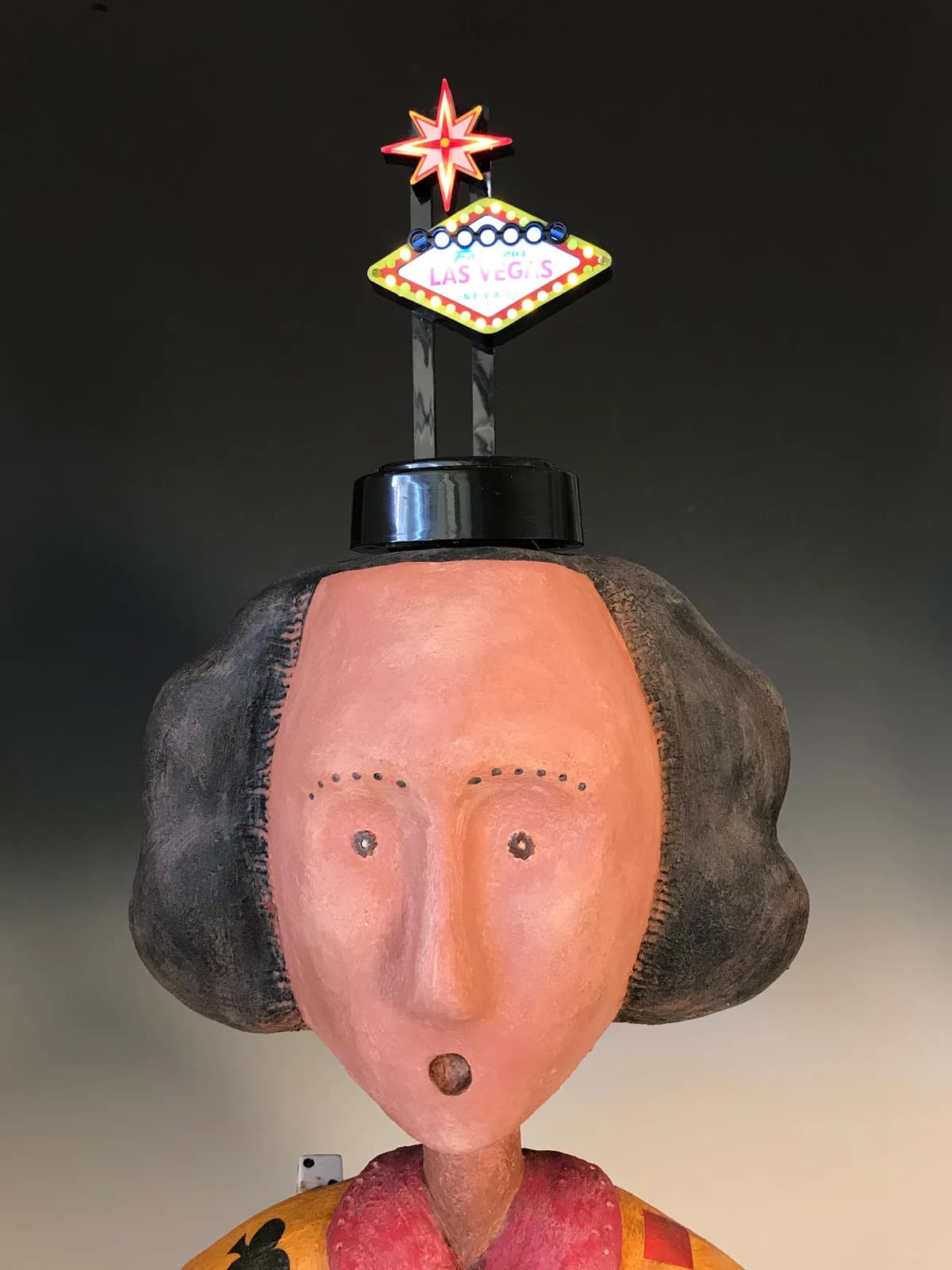 A folk art sculpture of a figure with a Las Vegas sign on its head, rendered in Japonica and Apple Blossom hues.