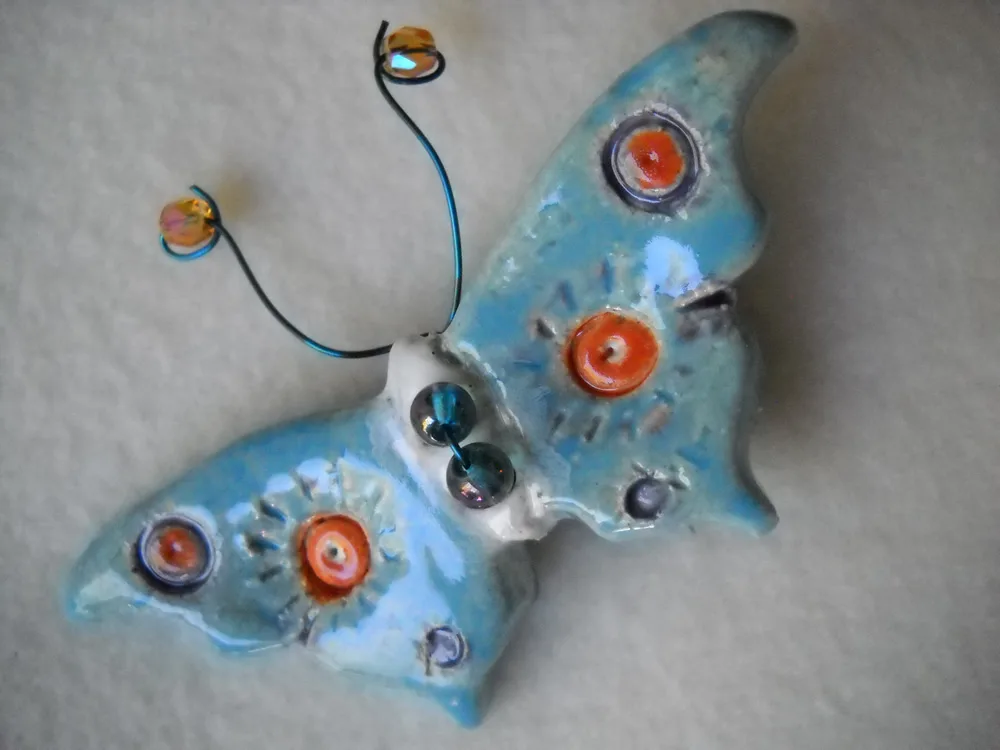 A whimsical ceramic butterfly sculpture with a blue glaze and orange accents, featuring wire antennae and beaded eyes.