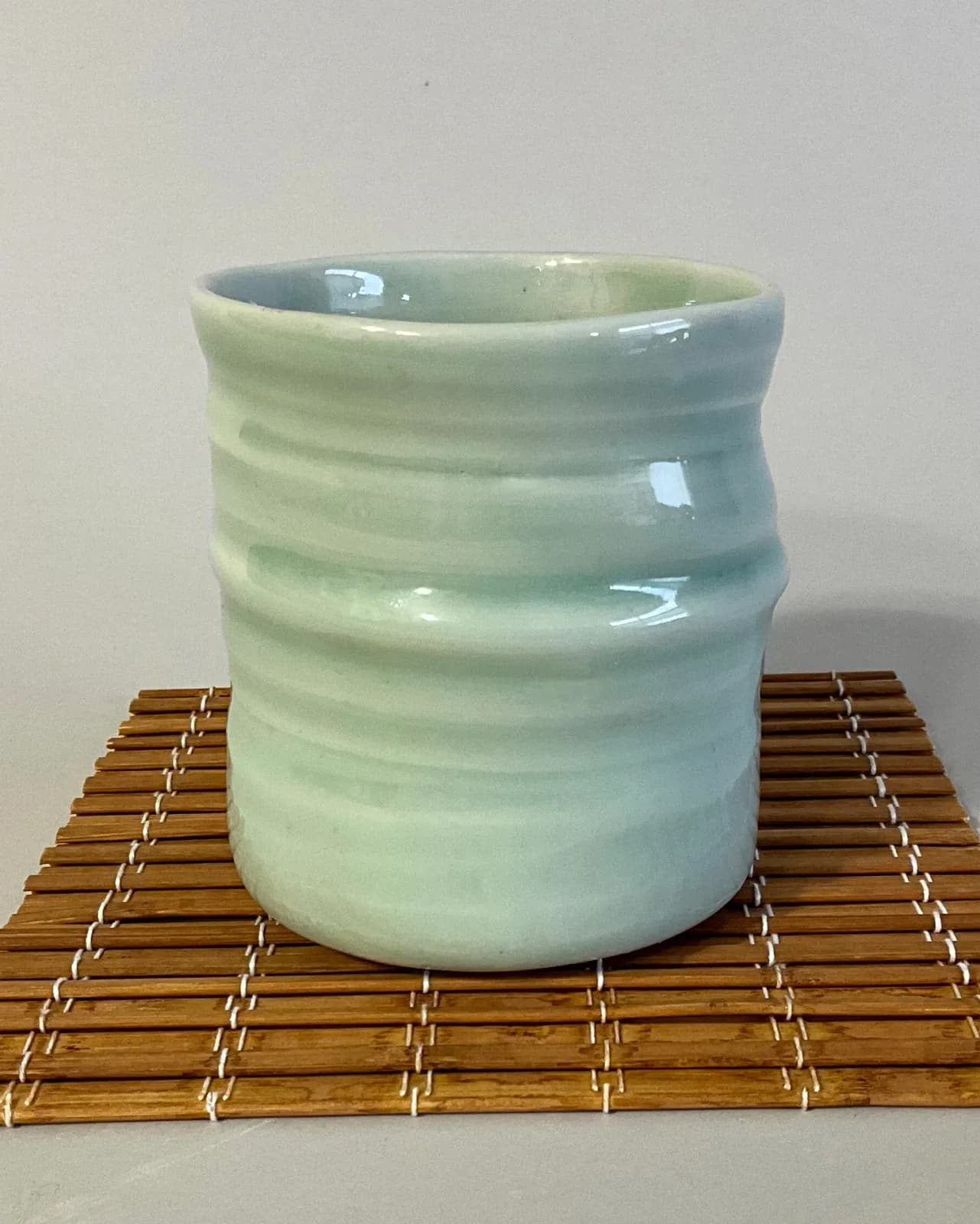 A celadon glazed ceramic cup with horizontal ribbing sits on a bamboo mat.