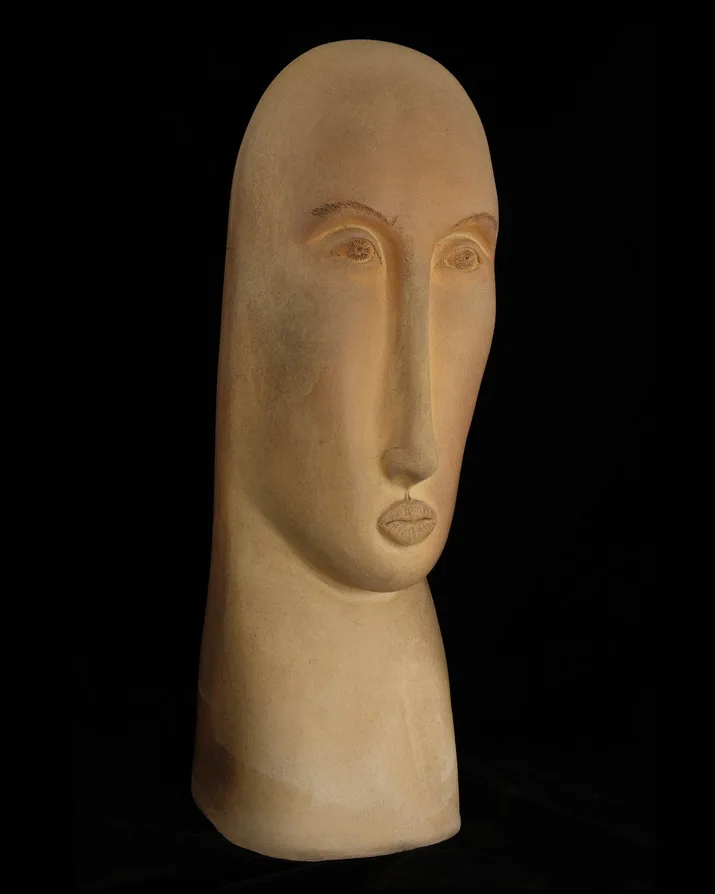 A clay bust of a stylized face with elongated features and a neutral expression against a black background.