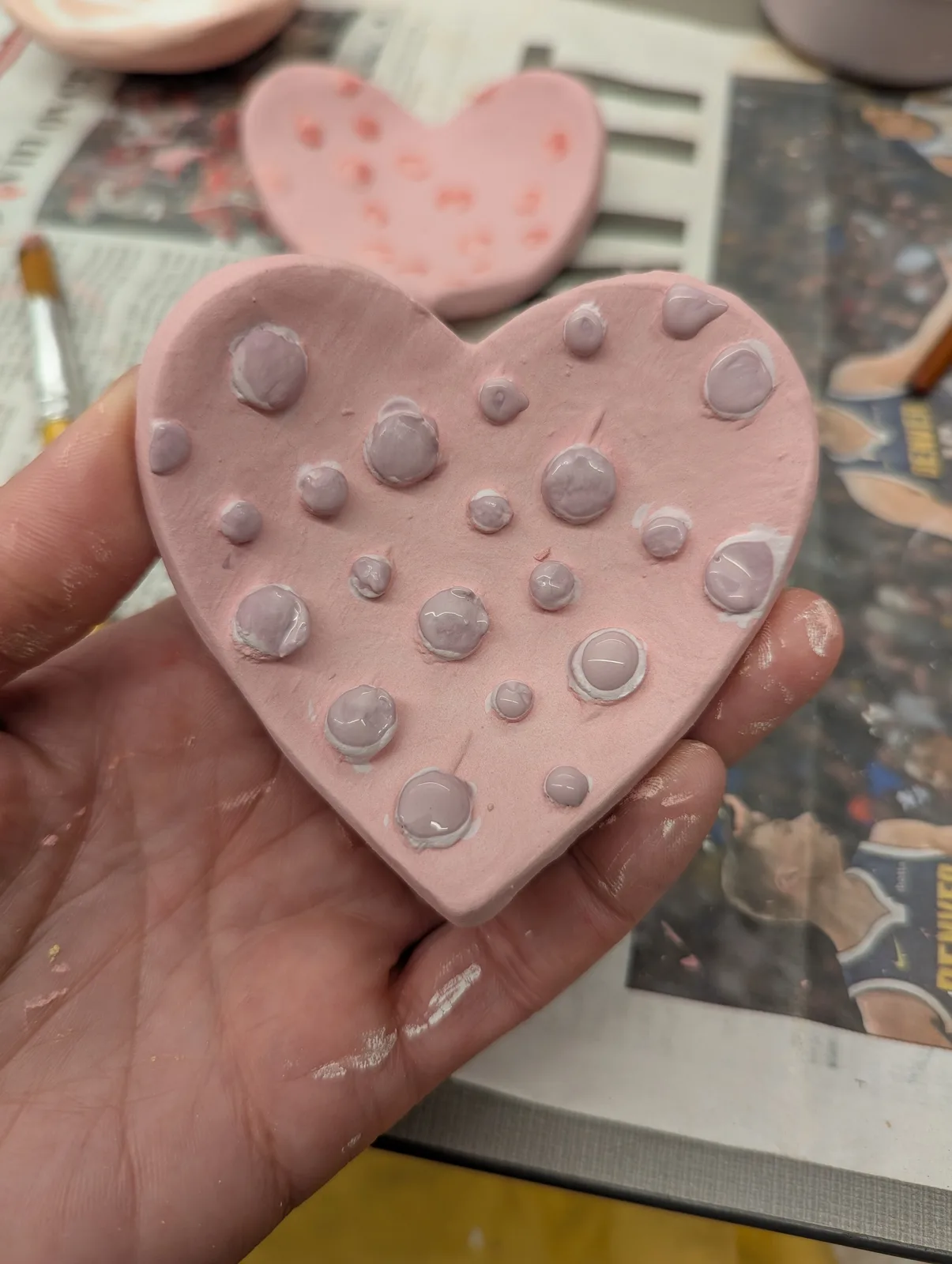 A pink heart-shaped ceramic piece with applied bubble-like forms is held in a hand, showing it in progress.