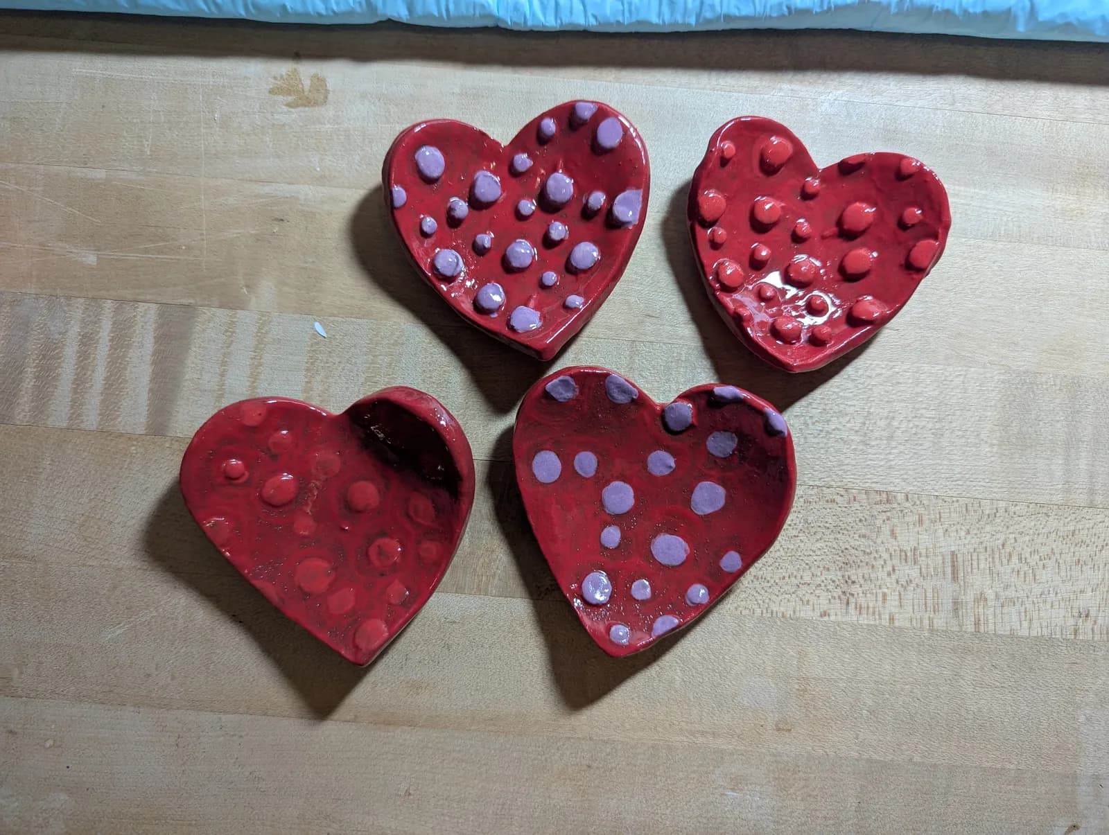 Four red ceramic heart-shaped dishes with pink polka dots are displayed on a wooden surface.